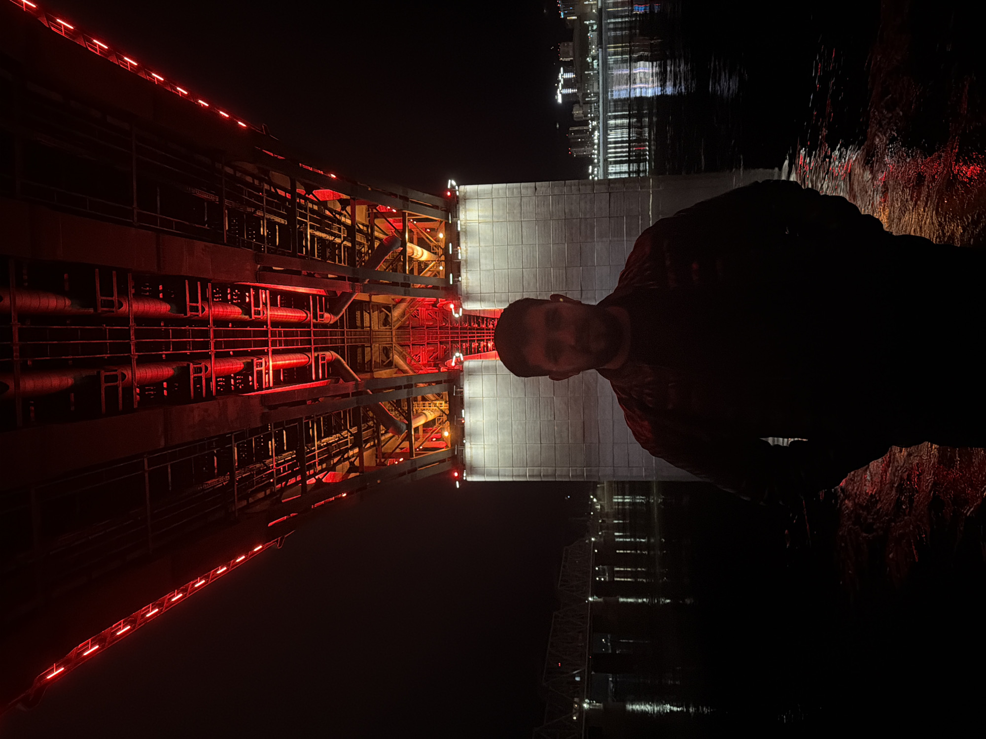 A nighttime shot shows a man standing near a bridge in Krasnoyarsk, Russia. The bridge is illuminated with red and white lights. In the background, city lights reflect on the surface of a body of water.