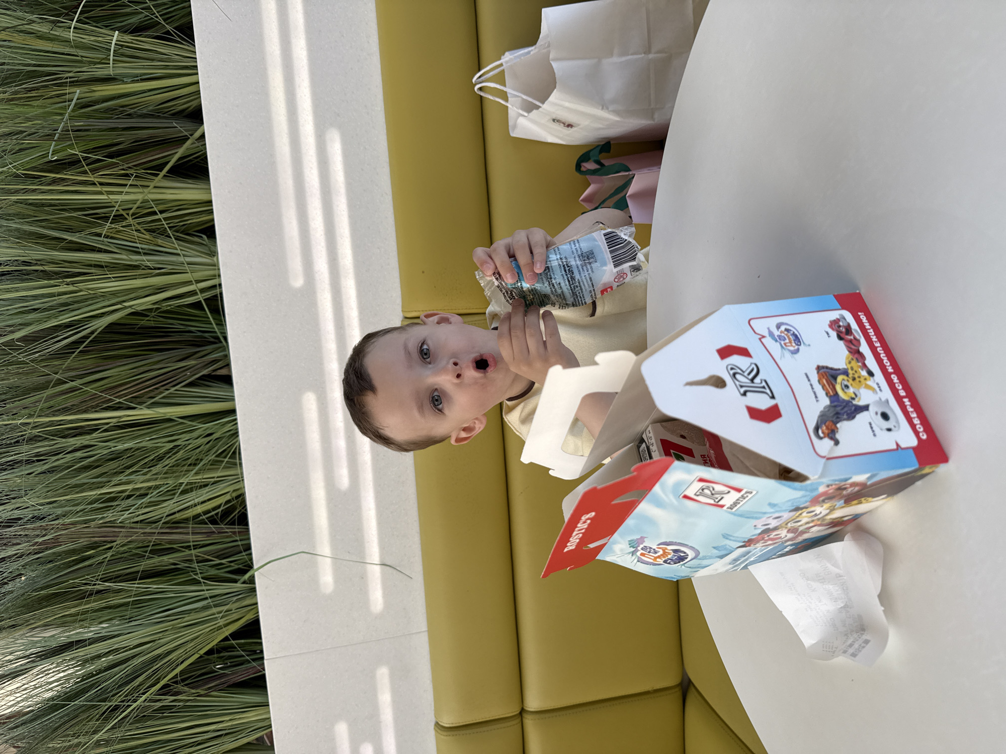 A young boy is sitting at a white table outside, holding a bottle and looking at the camera. A yellow cushioned bench serves as seating. On the table, next to the boy, is an open Rostic's chicken meal box featuring Paw Patrol characters. A white shopping bag sits behind him. To the left of the table is a wall lined with tall grasses. Text on the box indicates Russian and English languages. The location is likely Vladivostok, Russia, given the Rostic's chicken restaurant franchise is present in that city.