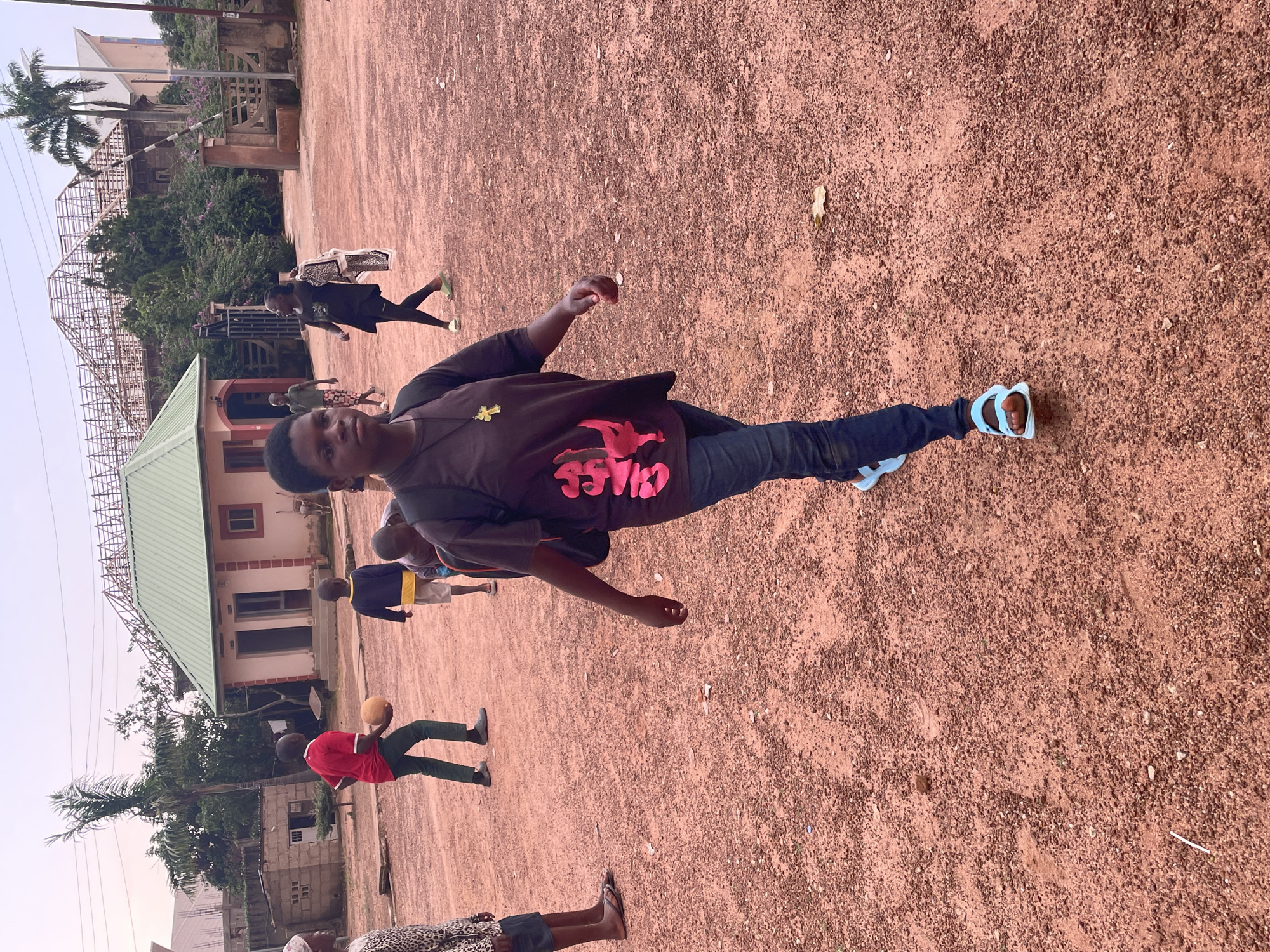 In Bar Jirgi Summa, Nigeria, a group of individuals are in an outdoor, dusty, reddish-brown area. In the foreground, a young person with short hair, wearing a dark t-shirt with a pink graphic and dark jeans, walks towards the camera, carrying a backpack. They are wearing light blue sandals. To their left, another person in a patterned top and dark shorts is also walking. Further back, a person in a red shirt and dark pants is holding an orange ball. A child in a blue and yellow shirt and light shorts is walking away from the camera. In the background, a light-colored building with a green roof is visible, along with other structures and trees under a hazy sky. The ground is covered in loose dirt and small pebbles.