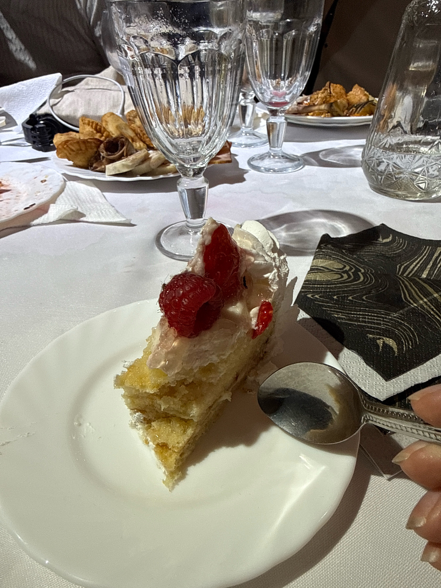 A slice of cake with raspberries and whipped cream sits on a white plate. The cake is layered with a yellow sponge and white frosting. A silver spoon rests on the plate, and a patterned napkin is to the right of the plate. In the background, there are two tall glasses, a glass decanter, and a plate of assorted appetizers. The scene is set on a white tablecloth.