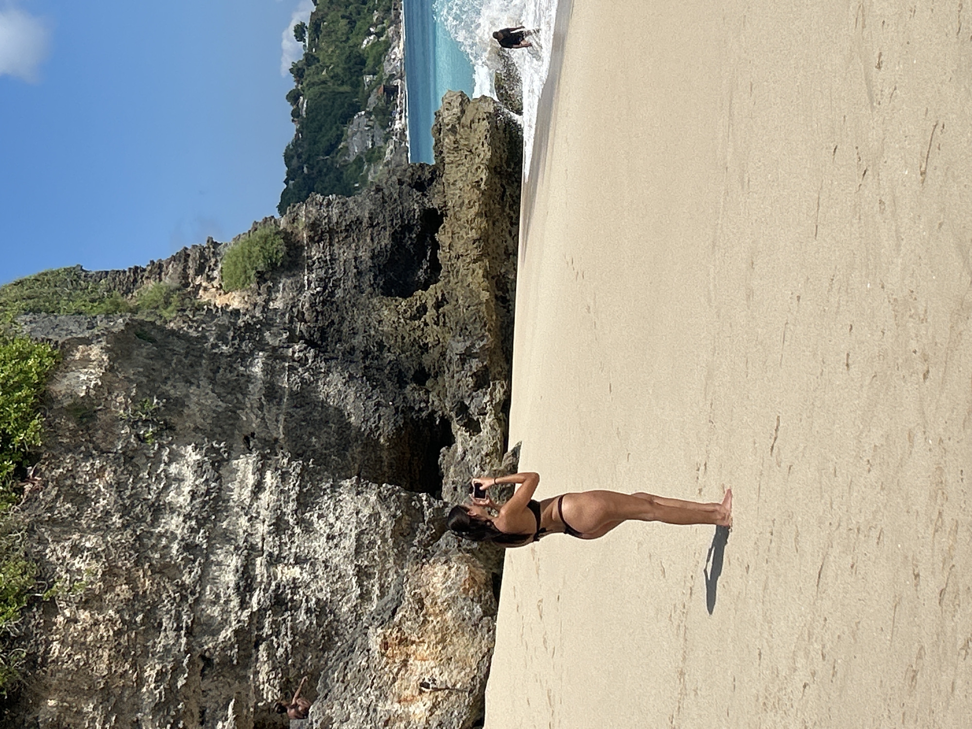 In Kuta Selatan, Indonesia, a woman in a black bikini stands on a sandy beach, holding a camera and taking a photo. She is facing away from the viewer, towards the ocean and a large rocky cliff face. The cliff is covered in greenery and rises steeply from the beach. Waves are crashing near the base of the cliff. In the distance, the ocean is a vibrant turquoise and a person is walking in the water. The sky is clear blue with a few scattered clouds. The sand is light tan and appears smooth with some light footprints visible.