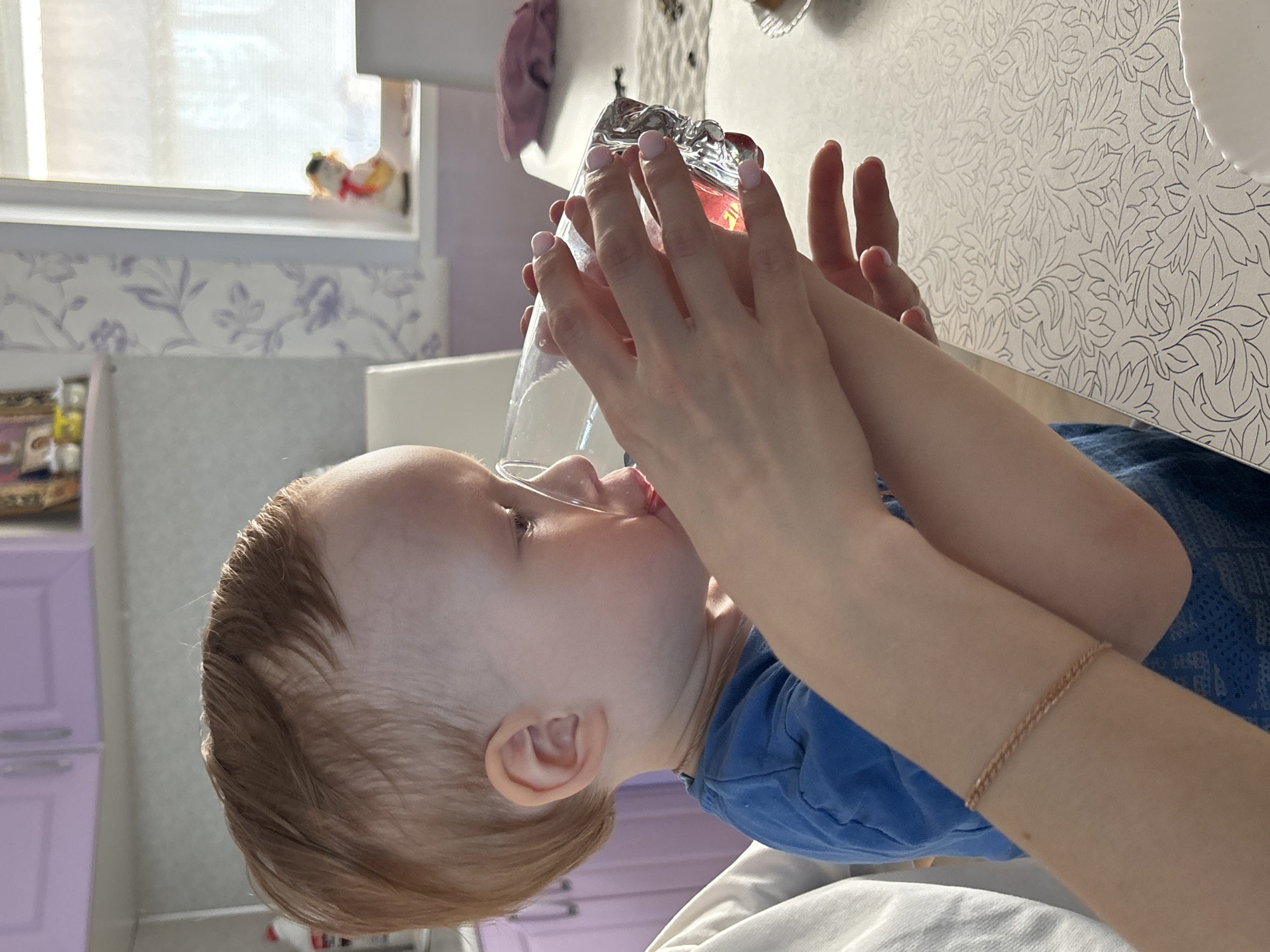 A toddler drinks from a glass held by an adult indoors in Ферма, Russia. The child, wearing a blue shirt, has light brown hair. The adult, wearing a gold-colored bracelet, supports the clear glass with both hands. The table has a white patterned surface. A window is visible in the background. The room is decorated with lavender-colored cabinets and floral wallpaper.