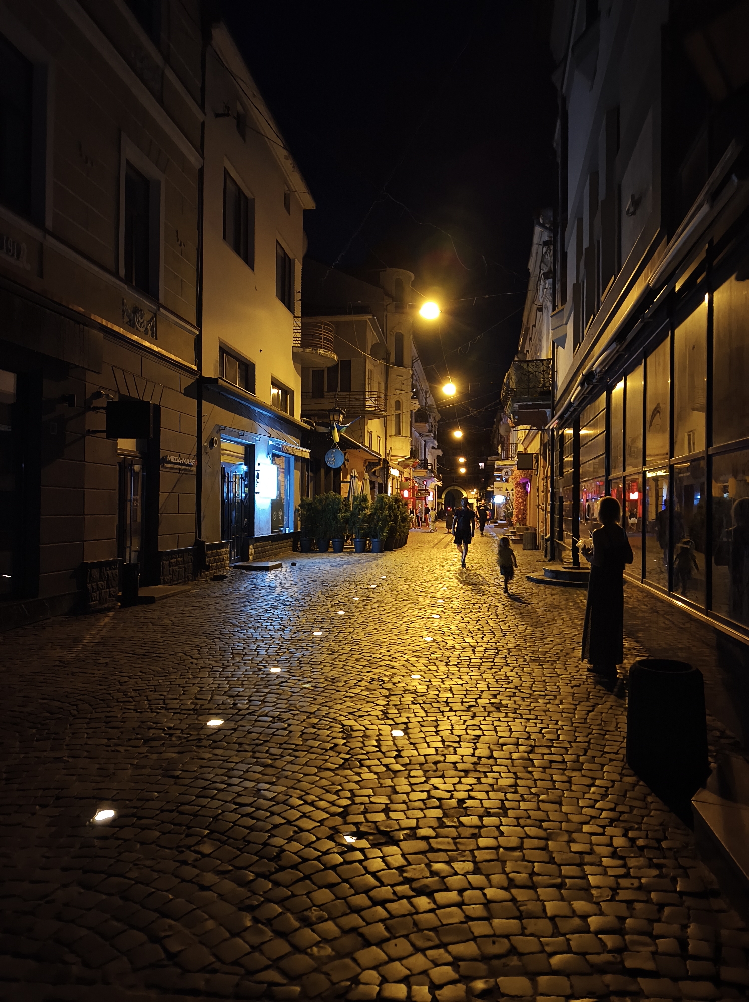 The image depicts a cobblestone street in the city of Ternopil, Ukraine, at night. The street is illuminated by streetlights and embedded lights in the pavement, casting a warm glow on the textured surface. On the left side of the street are buildings with windows and architectural details, some appearing to be shops or residences. On the right, a long building with large, reflective windows lines the street. Several people are visible: a woman standing in the foreground on the right, a man and a child walking away from the camera in the middle of the street, and other figures further down the street. The scene suggests a lively urban environment, even at night, with an inviting atmosphere created by the lighting and the architecture.