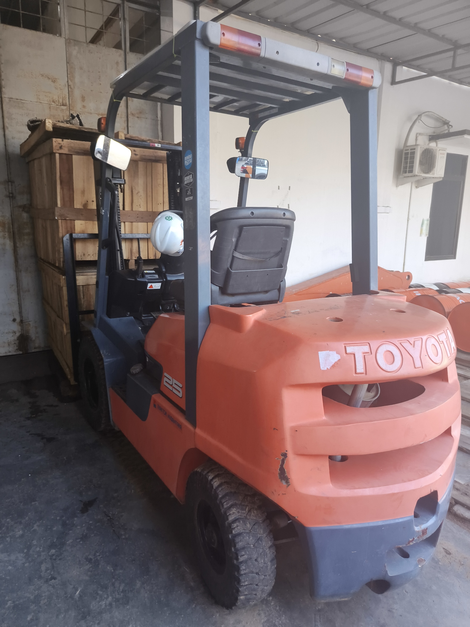 A Toyota forklift is parked indoors, with its front facing the viewer. The forklift is orange and gray, with a roll cage and a black seat. There are wooden crates stacked to the left of the forklift. In the background, there is an air conditioning unit mounted on the wall, and an exit door. The forklift appears to be in a warehouse or industrial setting. The scene is well-lit, with natural light entering from an unseen source. The ground is concrete, with some water stains. There are no people or animals visible in the image. The forklift is in good condition, with only minor scuffs and scratches visible. The image has a slightly grainy texture, and the colors are muted. The forklift is positioned at an angle, allowing a clear view of its front and side. The overall impression is one of a functional industrial space.