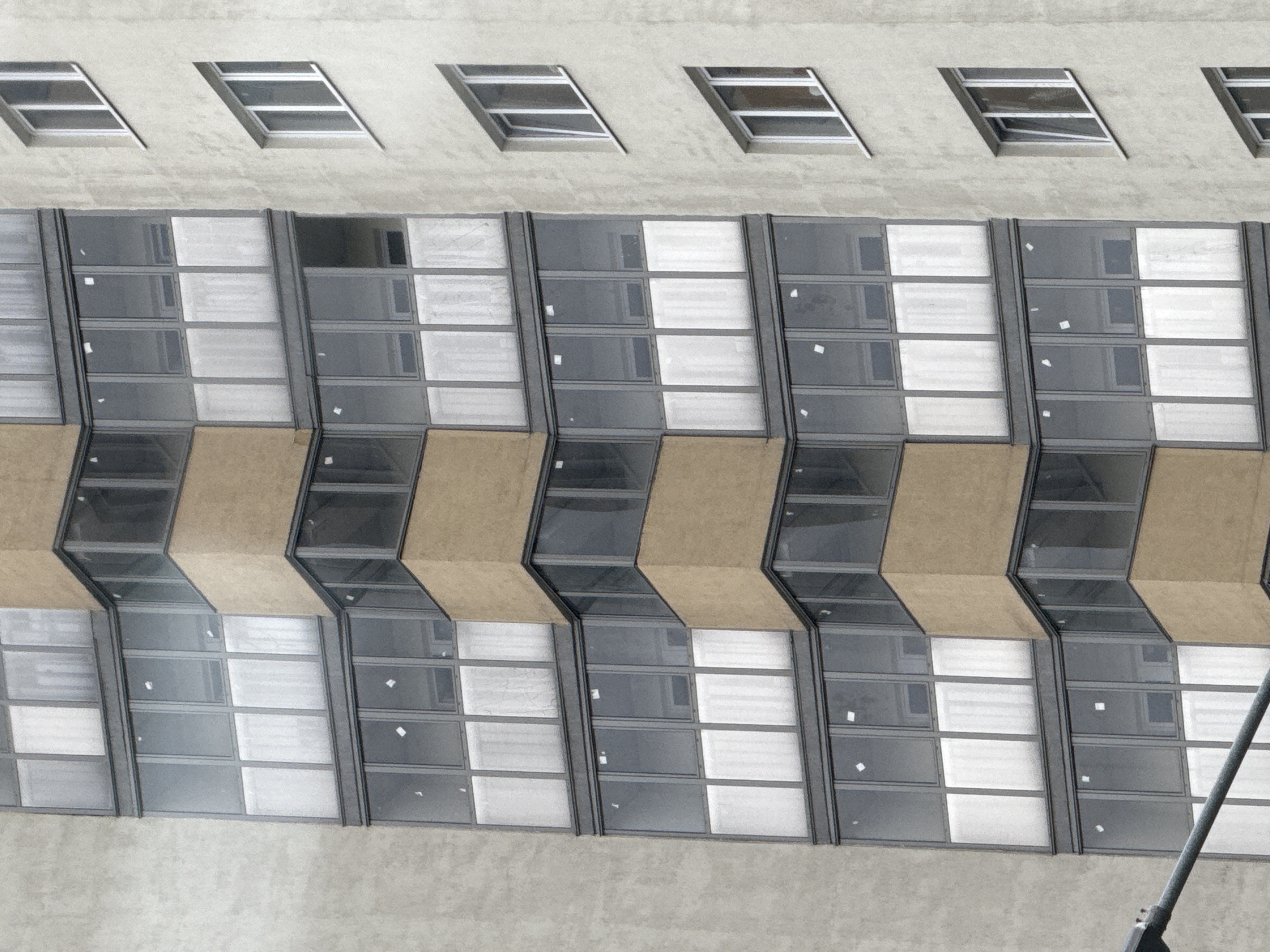 The image displays a section of a modern apartment building in Chelyabinsk, Russia. The building features a mix of concrete and beige-colored facade elements. The most prominent feature is the arrangement of windows, which are large and numerous.
The left side of the image shows a section of the building with a grey concrete facade. Adjacent to this is a protruding section with a beige facade. This beige section is adorned with a series of balconies or enclosed window units, characterized by their dark frames and glass panels. These panels are divided into multiple panes, some of which appear frosted or opaque, while others are transparent, revealing the interior spaces.
The right side of the image showcases another section of the building with a light grey concrete facade. This part of the building has a more traditional window arrangement, with several rectangular windows featuring white frames. These windows are spaced vertically and appear to be a mix of open and closed.
The overall impression is one of urban architecture, highlighting the residential nature of the structure. The lighting suggests it might be daytime, with diffused light, possibly indicating an overcast sky. No people are visible in the image. The environment is strictly architectural, focusing on the building's facade and its windows.