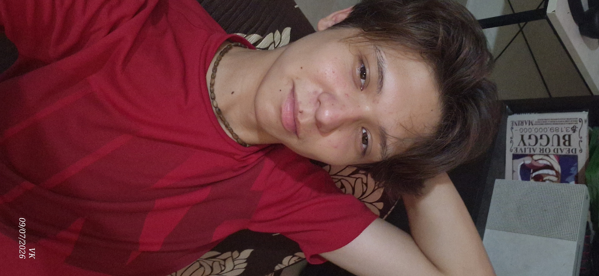 This is a portrait-oriented, close-up, selfie-style image of a young individual with medium-length, dark, brown hair. The individual has fair skin with some blemishes and is wearing a red t-shirt. A beaded necklace is visible around their neck. Their left arm is bent, with their hand resting behind their head, and their right arm is also bent, with their hand resting on their thigh or the surface they are sitting on. The background is out of focus but appears to include a shelf with some items, including what looks like a book or poster with the word "BUGGY" prominently displayed. The lighting is from the front, illuminating the subject's face. The image also has a watermark in the bottom right corner that reads "VK 09/07/2026". There is no indication in the image that it was taken in Vrindavan, India.
