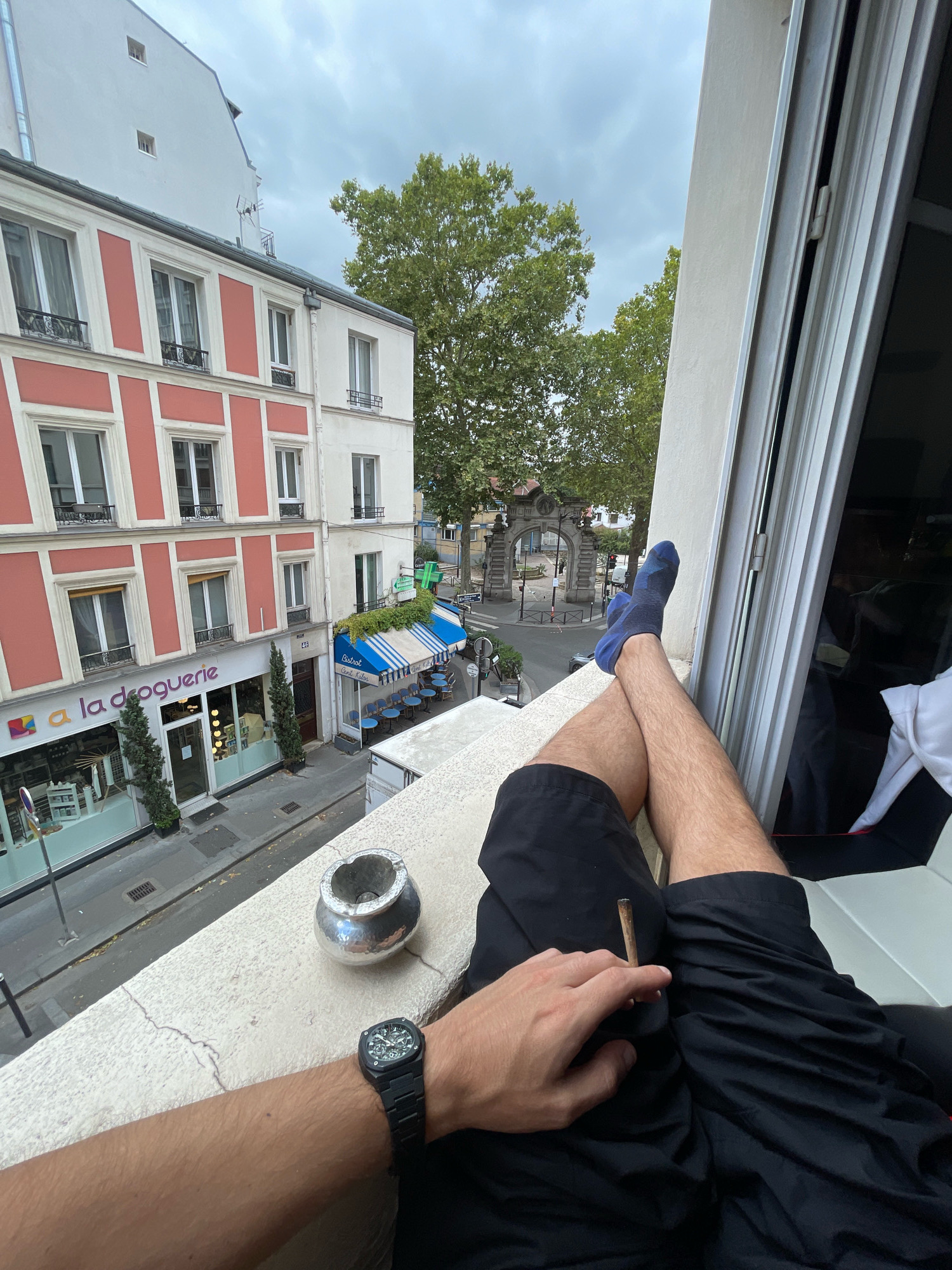 A first-person perspective captures a person relaxing on a balcony overlooking a street in Paris, France. The person's legs are crossed, wearing black shorts and blue socks. A lit cigarette is held in their right hand, and an ashtray sits on the balcony edge nearby. Below, the street features a building with a pink facade and white trim, housing a "la droguerie" store. Adjacent is a bistro with blue and white awnings and outdoor seating. In the background, a stone archway with decorative carvings stands at an intersection, and trees line the street. The sky is overcast, suggesting a cloudy day.
