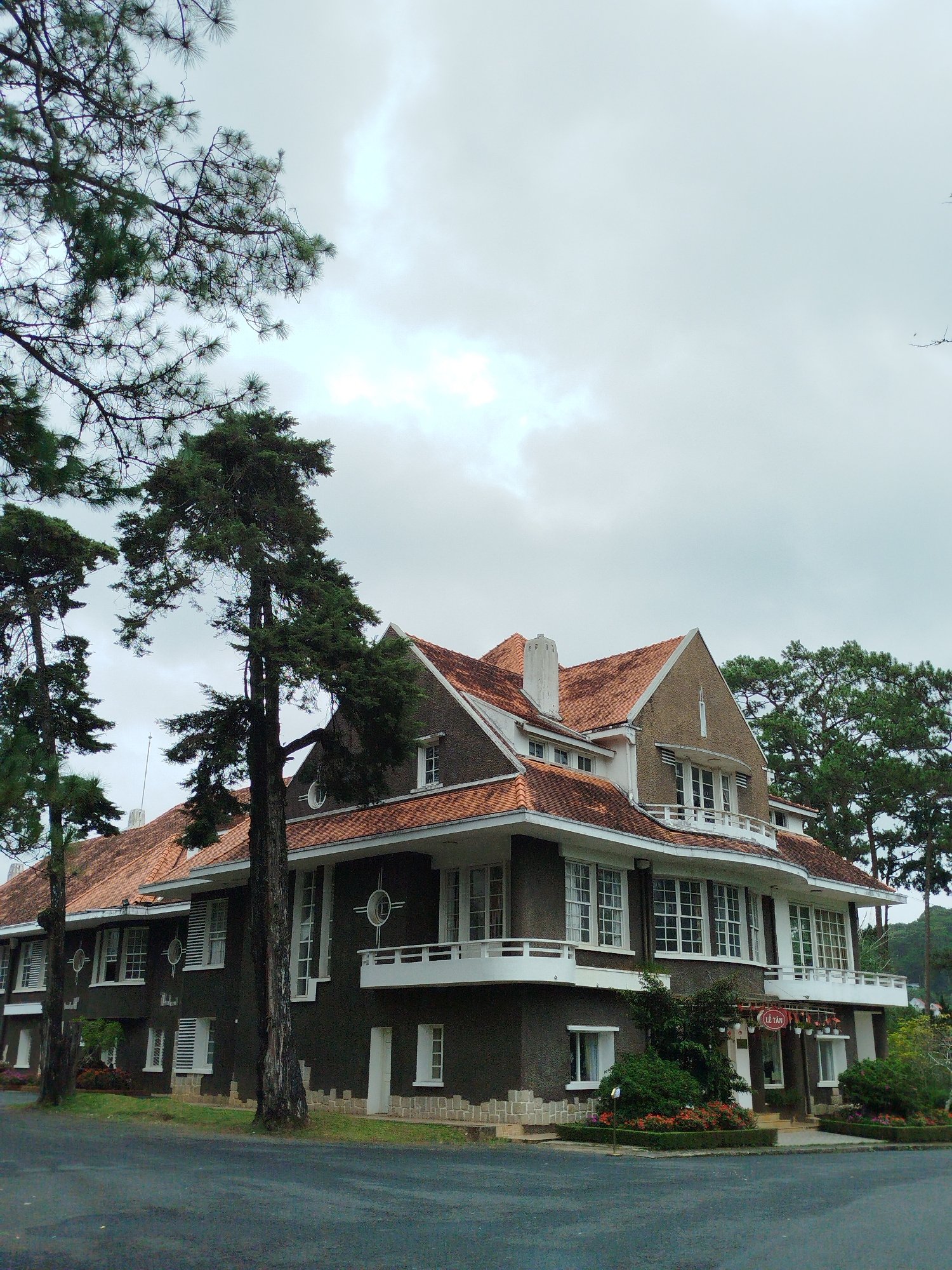 The image shows a large, dark-colored building with red-tiled roofs, characteristic of French colonial architecture in Da Lat, Vietnam. The building appears to be a hotel or guesthouse, with a sign that reads "LE TAN" above the entrance. There are many windows, some with white shutters, and balconies on the upper floors. The building is surrounded by lush greenery, including tall pine trees and manicured gardens with red flowers. The sky is overcast, suggesting a cloudy or misty day. The overall impression is one of a historic and somewhat secluded property, likely a tourist attraction or accommodation in the mountainous city of Da Lat.
