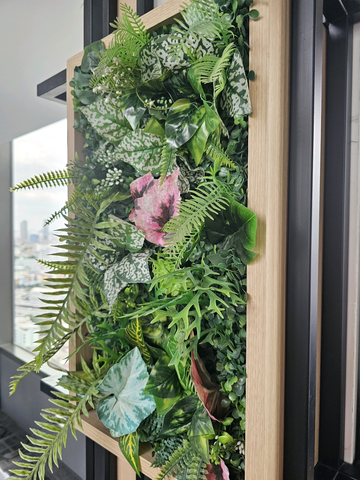 This is a close-up, eye-level, outdoor shot of a vertical garden wall composed of various artificial plants, including ferns, broad-leafed plants, and smaller foliage with white speckles, and a single pink flower with dark markings. The garden is housed within a light-colored wooden frame. The background reveals a blurred view of a cityscape, suggesting an urban environment. Specifically, the cityscape visible through the window in the background, with its characteristic mix of modern and older buildings, is consistent with the city of Bangkok, Thailand.