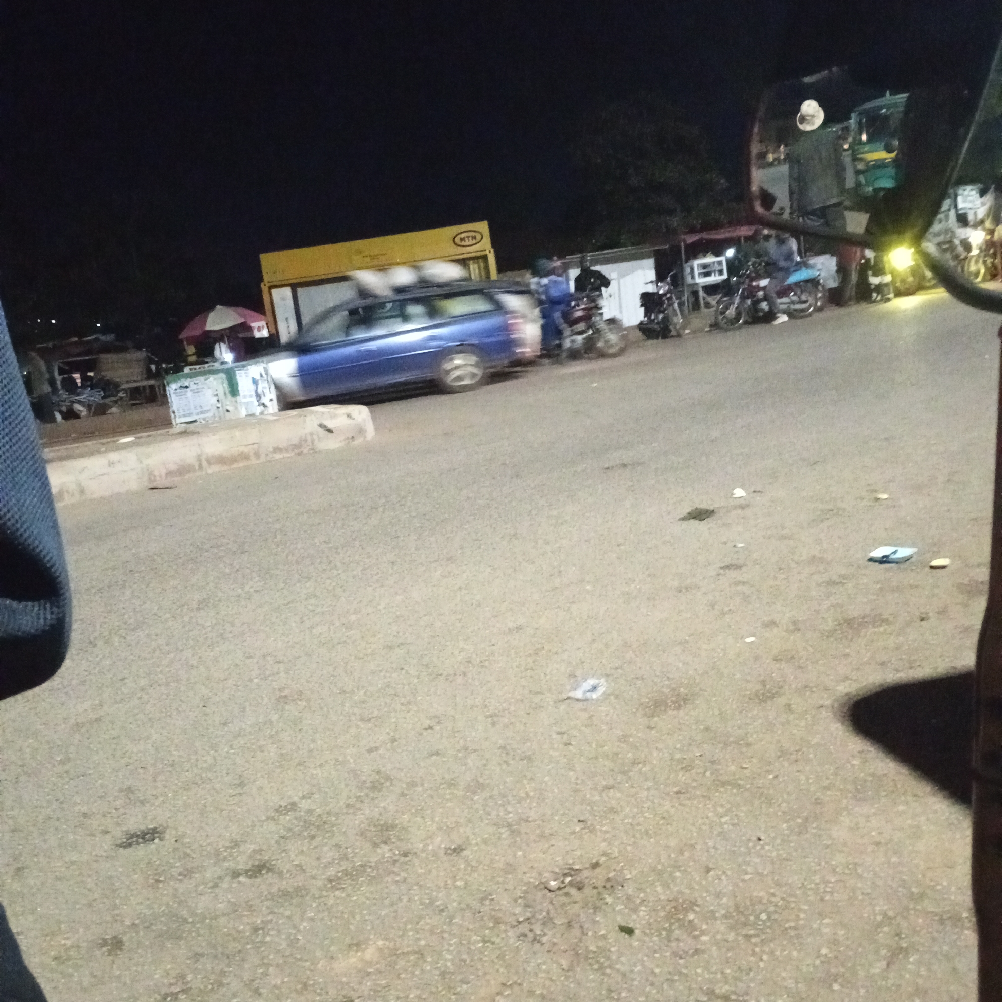 At night in Bukuru, Nigeria, a blue station wagon is parked on a paved road. Beside the car, a man in a blue shirt and white cap is seated on a motorcycle. In the background, a yellow building with the logo "MTN" is visible. Several other motorcycles are parked nearby, and a person in a black jacket stands near one of them. The scene suggests a roadside area with commercial activity.
