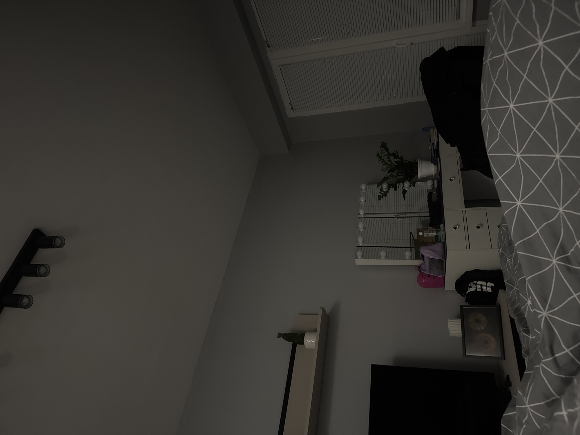 A bedroom is visible. A bed with a grey geometric patterned duvet is in the right portion of the frame. A white vanity with drawers and mirrors is positioned near the bed. A black backpack and a framed picture are placed on the vanity surface. A television and a white shelf holding a small plant are positioned on the wall below the vanity. A ceiling light fixture is in the upper center of the frame. A black light fixture is visible on the left wall. A plant is on top of the vanity. The location is an interior residential space and likely in Sofia, Bulgaria.