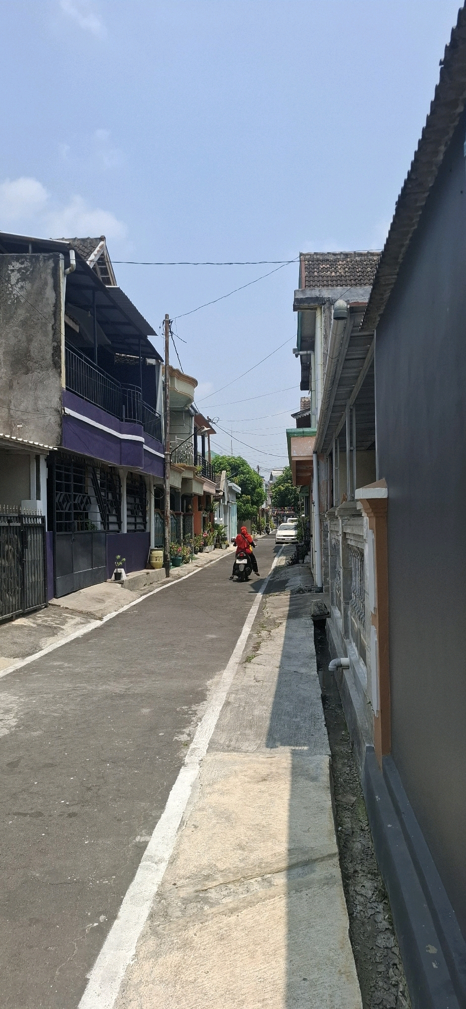 The image depicts a residential street in Desa Gagaksipat, Indonesia. The street is paved and has a white line marking the edge of the roadway. On the left side of the street are multi-story houses with balconies and dark-colored gates. The house closest to the foreground has a purple lower section and a grey upper section. On the right side of the street, there is a single-story building with a dark grey facade and decorative patterned walls. A narrow, open drainage ditch runs alongside the sidewalk on the right. In the middle of the street, a person wearing a red headscarf and a red top is riding a scooter, moving away from the camera. A white car is parked further down the street on the right side. The sky is clear and blue, suggesting a sunny day. The environment appears to be a typical neighborhood setting with residential buildings and a paved road.