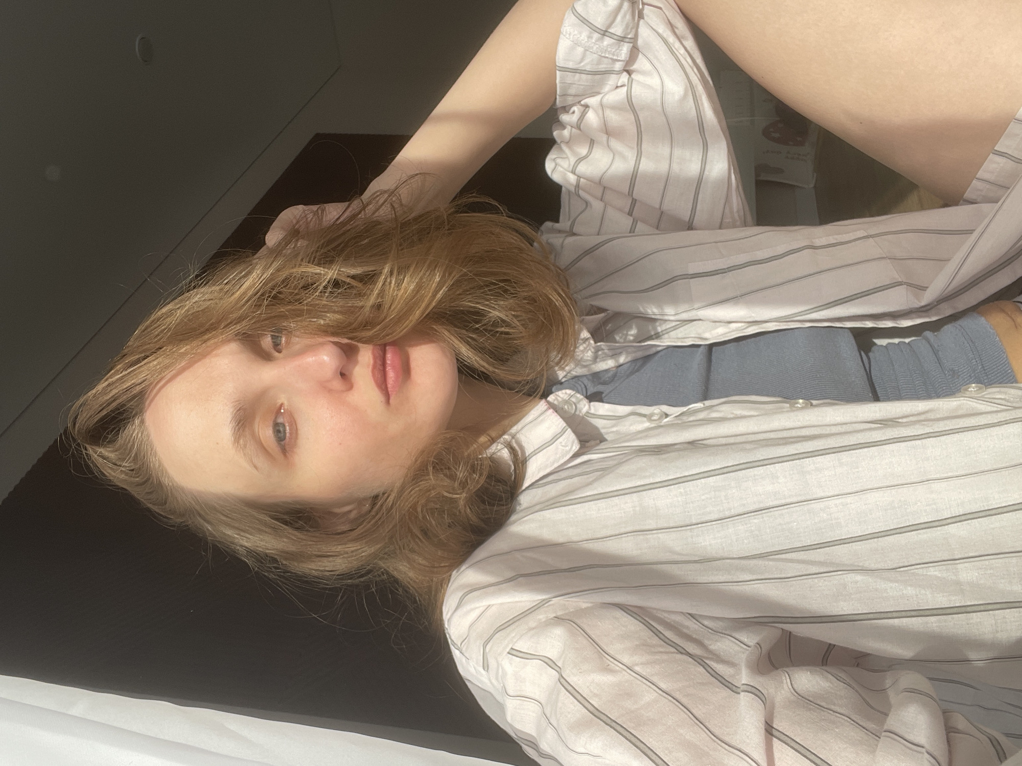 A woman lies on a dark surface, possibly a bed or floor, in an interior space. She is facing the camera with her head tilted. She wears a striped, light-colored, long-sleeved shirt over a blue garment. Her hair is blonde and wavy, partially obscuring her face. The lighting is uneven, with strong shadows cast on the left side of the frame and brighter illumination on the right. The background is blurred, but suggests a room with varying levels of light and dark. There is no information about the location in the city of Sepych, Russia.