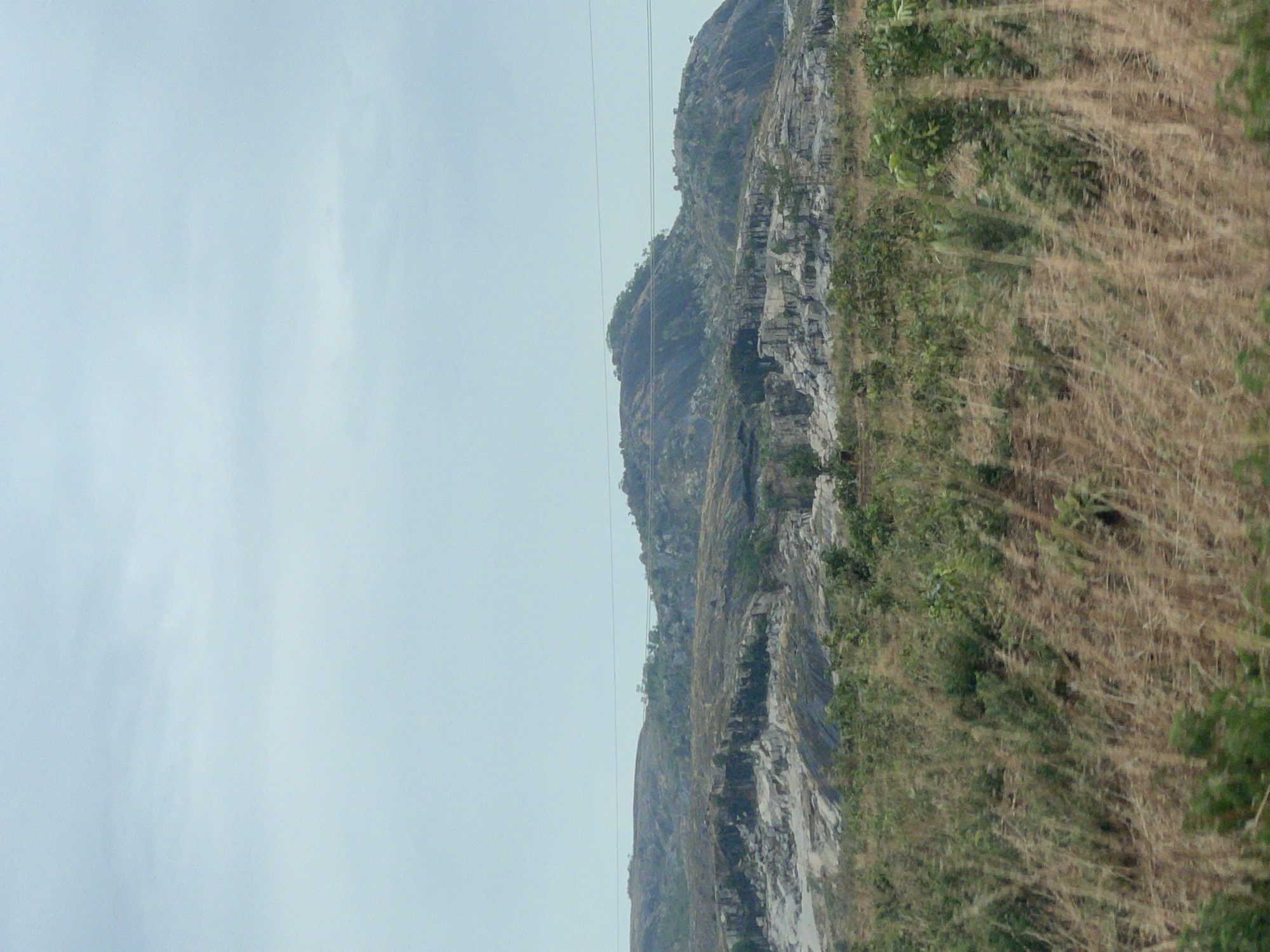 The image depicts a mountain near Kujama, Nigeria. The mountain face is rocky and layered, with sparse vegetation growing on its slopes. Dry, tall grass occupies the foreground. Overhead power lines are visible against the overcast sky.