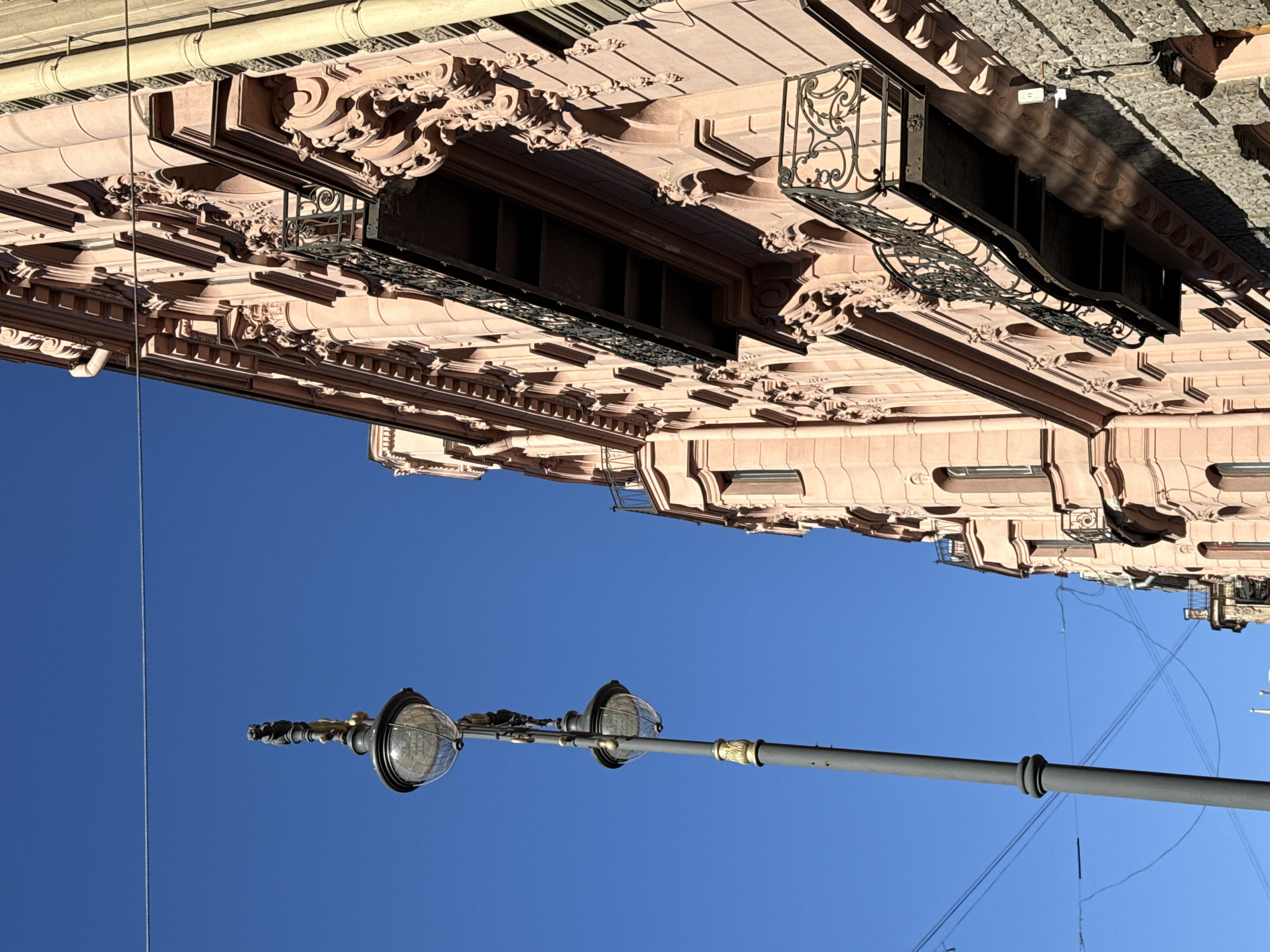 The image depicts a building facade and a lamppost against a clear blue sky. The building features ornate architectural details, including moldings, window frames, and decorative wrought-iron balconies. The building is light brown or beige in color. The lamppost is gray, with two round lamps affixed to a horizontal pole. There are several power lines. The location is likely Saint Petersburg, Russia, given the architectural style of the building and the style of the lamppost.