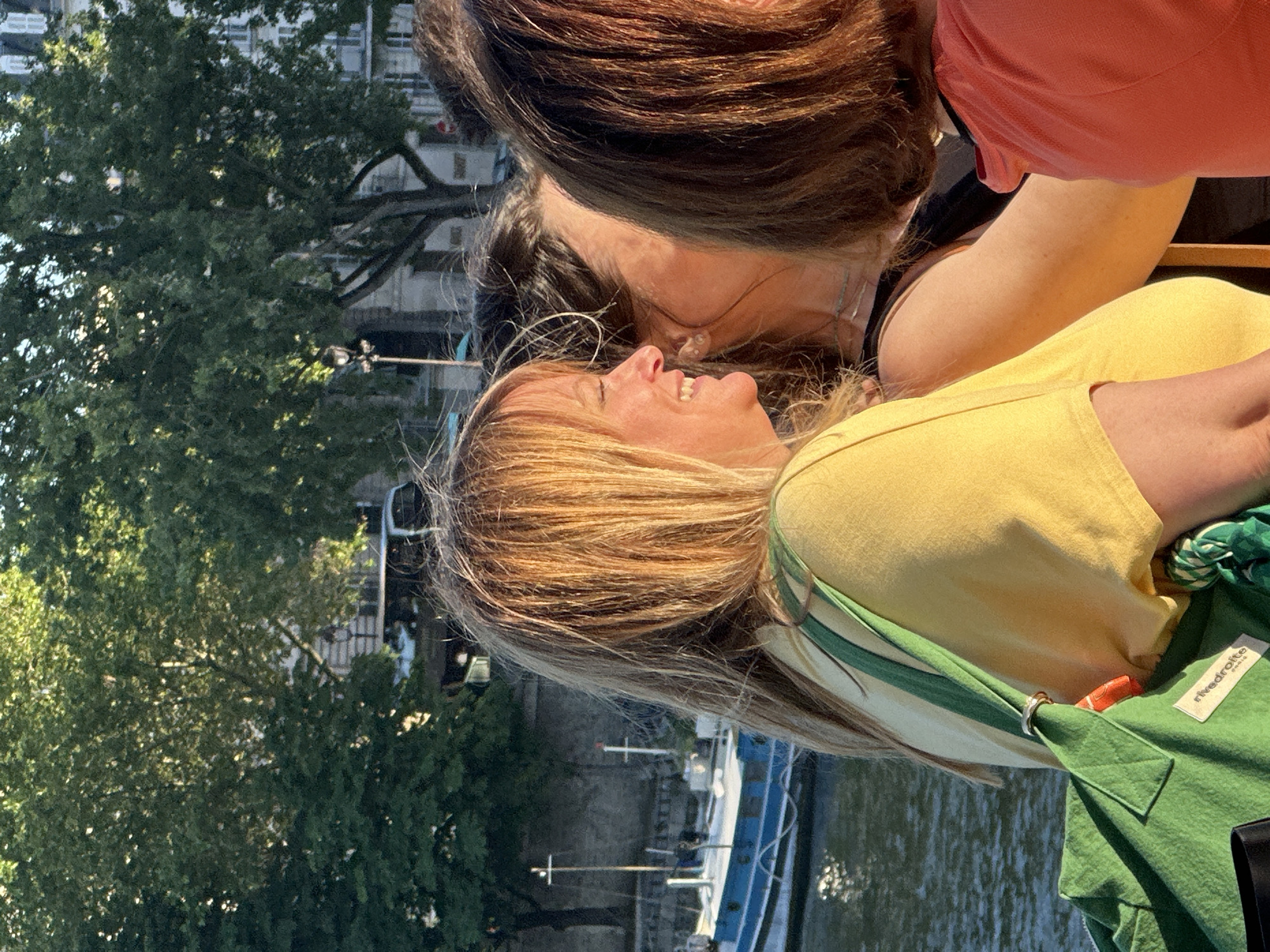 Two women are intimately embracing near a canal. One woman with blonde-streaked hair, wearing a yellow top and a green bag, smiles broadly. The other woman, with dark brown hair and wearing a red top, leans in close. In the background, there's a canal with docked boats and lush green trees. Buildings line the background with visible street lighting. The location is suggested to be Paris, France, given the architecture and the canal-side setting.