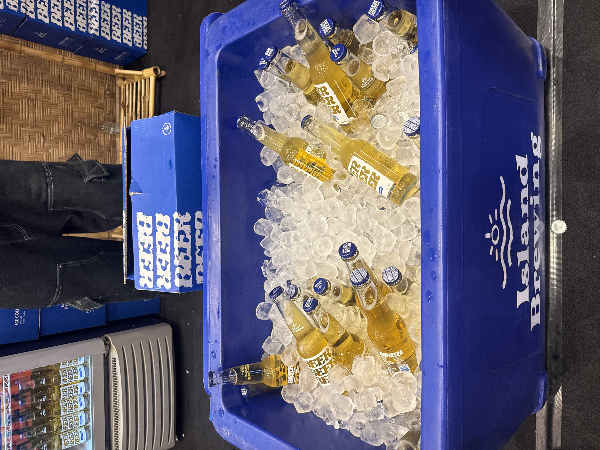 An overhead shot depicts a vibrant scene centered around a large, blue plastic cooler filled with ice and numerous bottles of beer. The golden-amber beer bottles are partially submerged in the ice, their blue and white caps bearing the text "FRESH BEER THINGS." Many labels on the bottles prominently feature "BEER" repeated in white text and the brand "Island Brewing," with "330 ML" also visible.
To the left of the cooler, several blue cardboard boxes are stacked, also branded with repeated white "BEER" text. A person's lower body, clad in black jeans, is partially visible standing near these boxes. Further left and lower, a commercial glass-front refrigerator is seen, fully stocked with more beer bottles, some labels displaying "ICE COLD" and "4.9% ABV."
The setting features a textured bamboo mat as flooring, and a small bamboo stool or stand is visible in the background. The side of the blue cooler clearly shows the "Island Brewing" logo and text. The scene appears to be a beverage sales or service point, likely indoors or under a cover, in a location like Kuta Selatan, Indonesia, as suggested by the local brand and aesthetic cues.