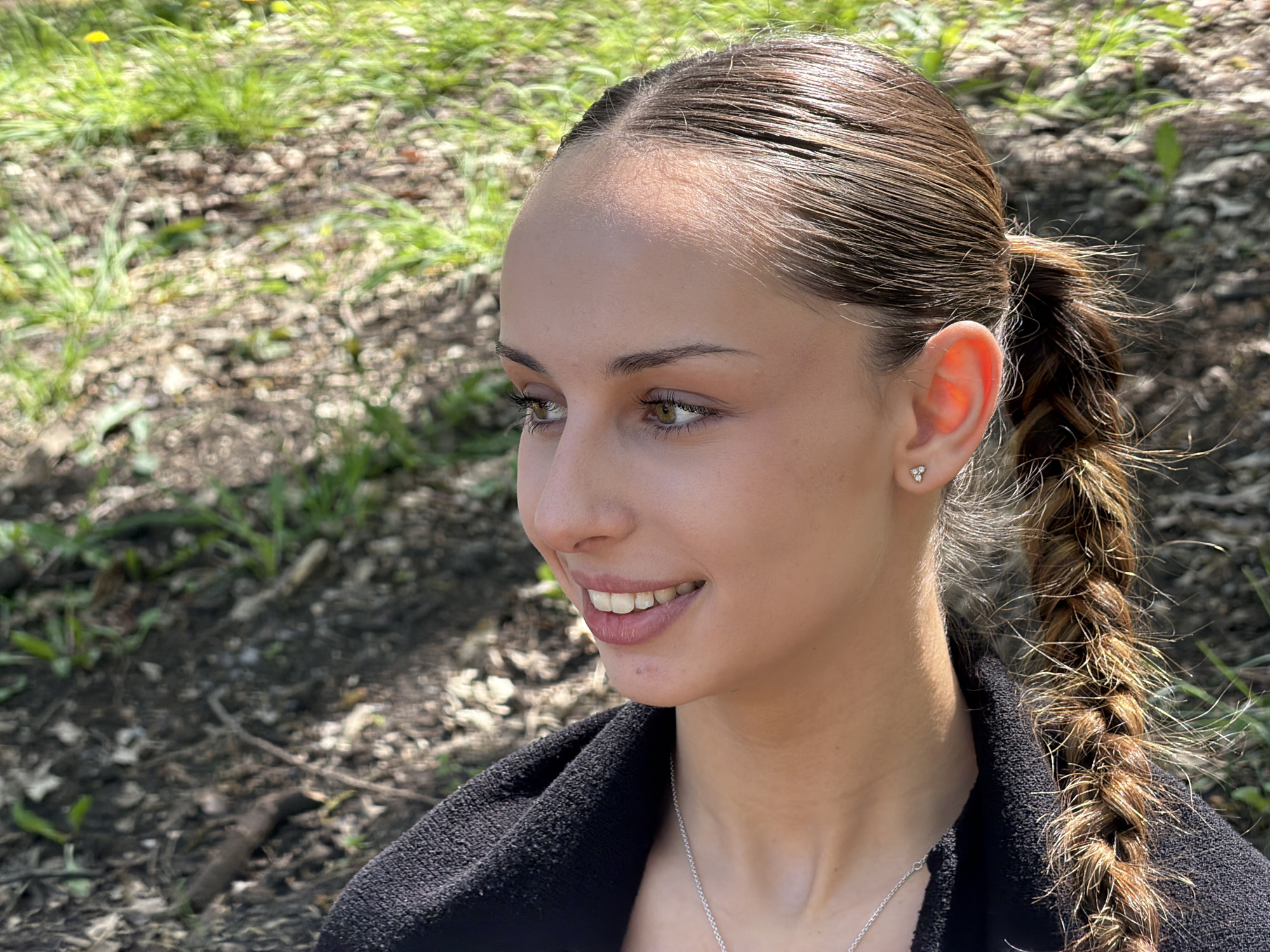 A young woman is outdoors on a sunny day. She has her hair pulled back in a tight braid, with loose strands near her face and braid. She is wearing a black top and a silver necklace. She is smiling and looking to her left. The background is blurred, but appears to be a grassy area with some foliage. The location is unknown.
