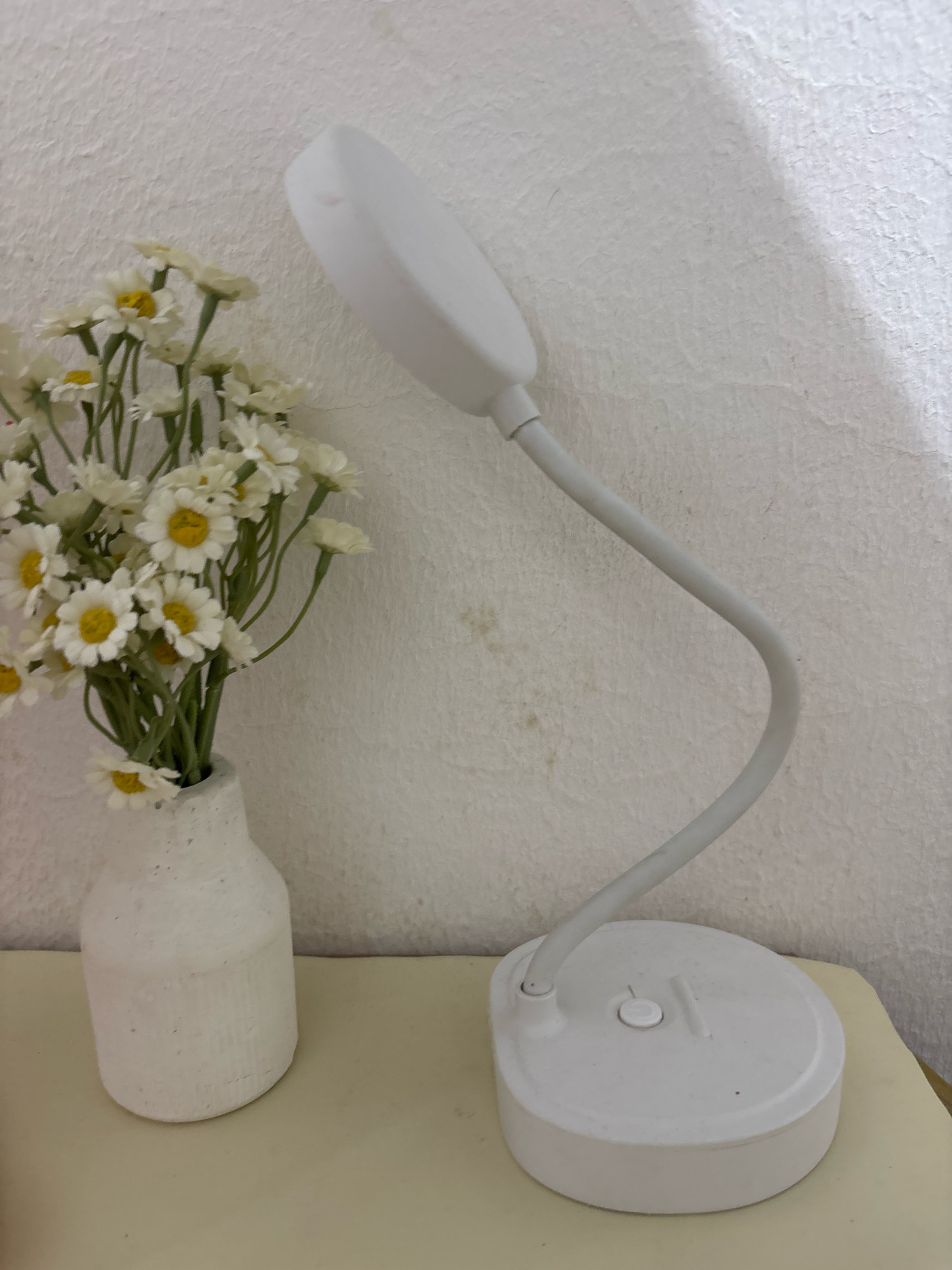 A white desk lamp with a flexible gooseneck arm is positioned on a light-colored surface to the right of a white vase filled with small white flowers. The lamp has a round base with a button and another indicator on its surface. The vase is textured and appears to be made of a rough material. The background is a white, textured wall. The scene is indoors, likely on a desk or table, in a residential setting in Wanajaya, Indonesia.