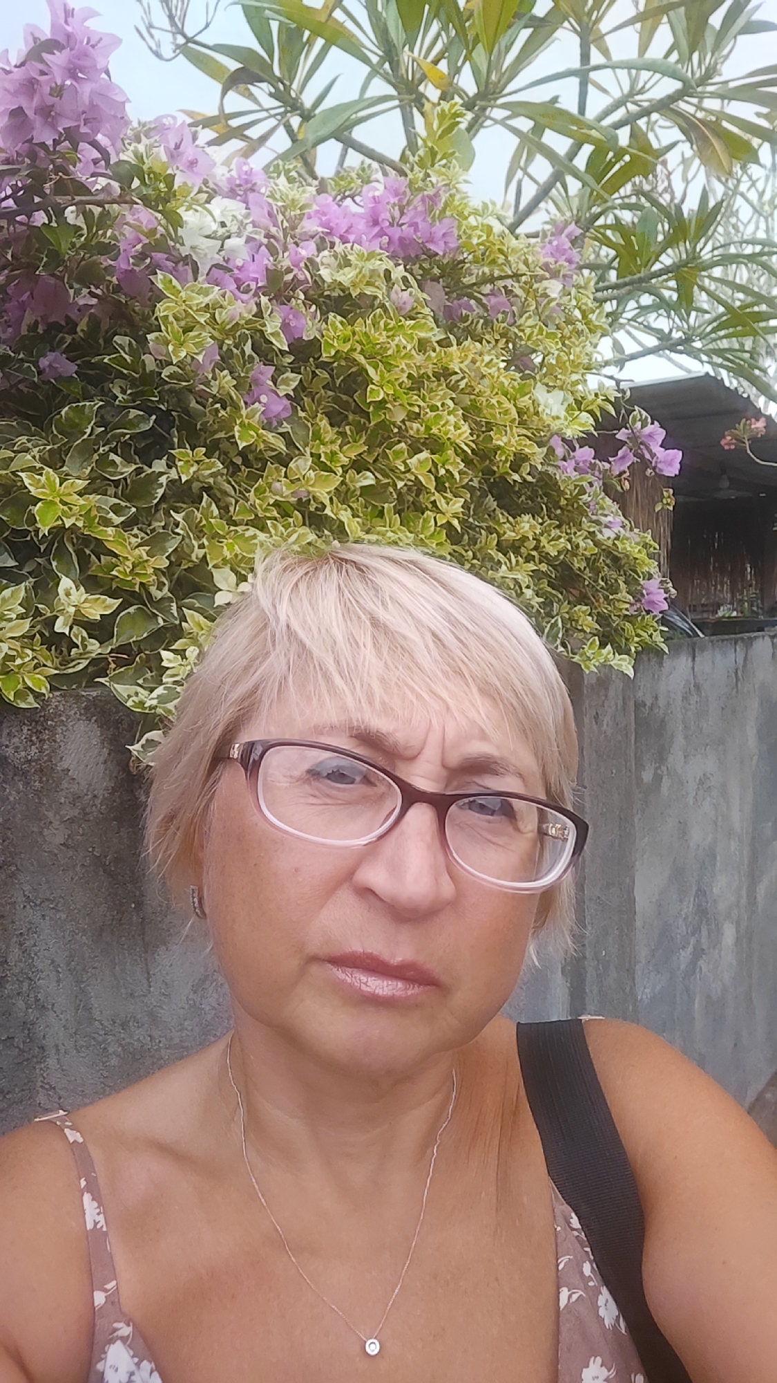 A woman with short blonde hair and glasses stands in front of a concrete wall adorned with flowering bushes featuring light purple flowers and variegated green and yellow leaves. Behind the wall, glimpses of vegetation and a roof suggest an urban setting. The woman wears a floral-patterned top and a necklace with a circular pendant. A dark-colored strap is visible over her shoulder. The scene appears to be in Nusa Penida, Indonesia, based on the location request.