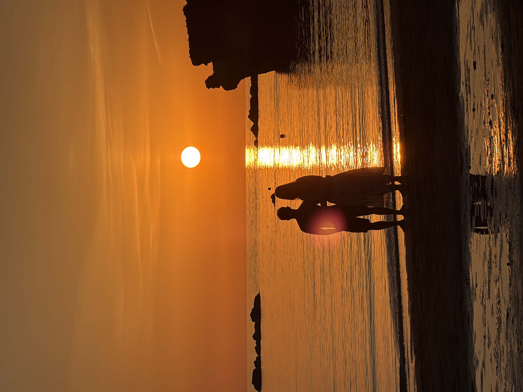 In Anzio, Italy, two figures, silhouetted against a brilliant orange sunset, stand on a sandy beach by the Tyrrhenian Sea. The sun, a bright disc, is low in the sky, casting a shimmering path of light across the water. The silhouettes, one appearing to be a male with shorts and the other a female in a flowing dress, stand close together, facing the sea. Several dark rocks are visible in the water, and a rocky cliff formation dominates the right side of the frame. The tide is out, leaving shallow pools of water on the wet sand, reflecting the warm hues of the sunset.