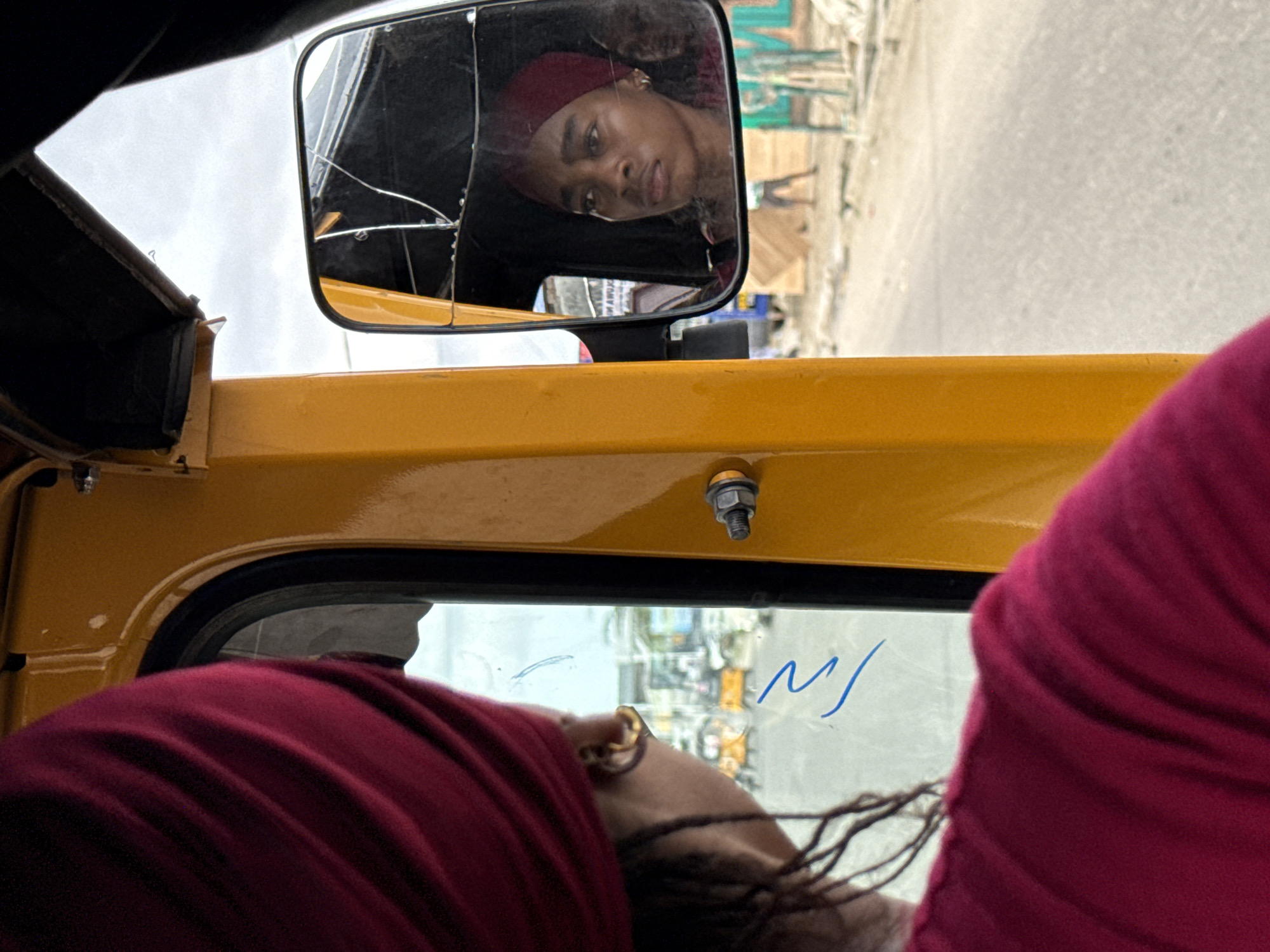 The image captures a scene from inside a yellow three-wheeled vehicle, possibly a rickshaw or 'keke napep', during the daytime under an overcast sky.
In the foreground, the back of a person's head and shoulder, covered in a deep red fabric, occupies the bottom right corner of the frame. An earring is visible on their ear, and their hair appears to be in braids or dreadlocks. Abstract blue markings, resembling a stylized "M/" or similar, are scrawled on the vehicle's window glass beside this person.
The main subject is a young woman reflected in a rectangular rearview mirror, positioned towards the top left. The mirror glass is visibly cracked in several places. The woman, who appears to be of African descent, is wearing a deep red head covering, matching the fabric in the foreground, and looks into the mirror with a neutral or slightly pensive expression. Her face is in clear focus.
The vehicle's interior frame is a bright yellow-orange. Outside the window and visible in the mirror's background, a blurry street or road is seen, lined with indistinct buildings and structures, indicating an urban or semi-urban setting. The blurriness suggests the vehicle might be in motion. The scene depicts a moment of travel or daily life in what is likely Ado, Nigeria.