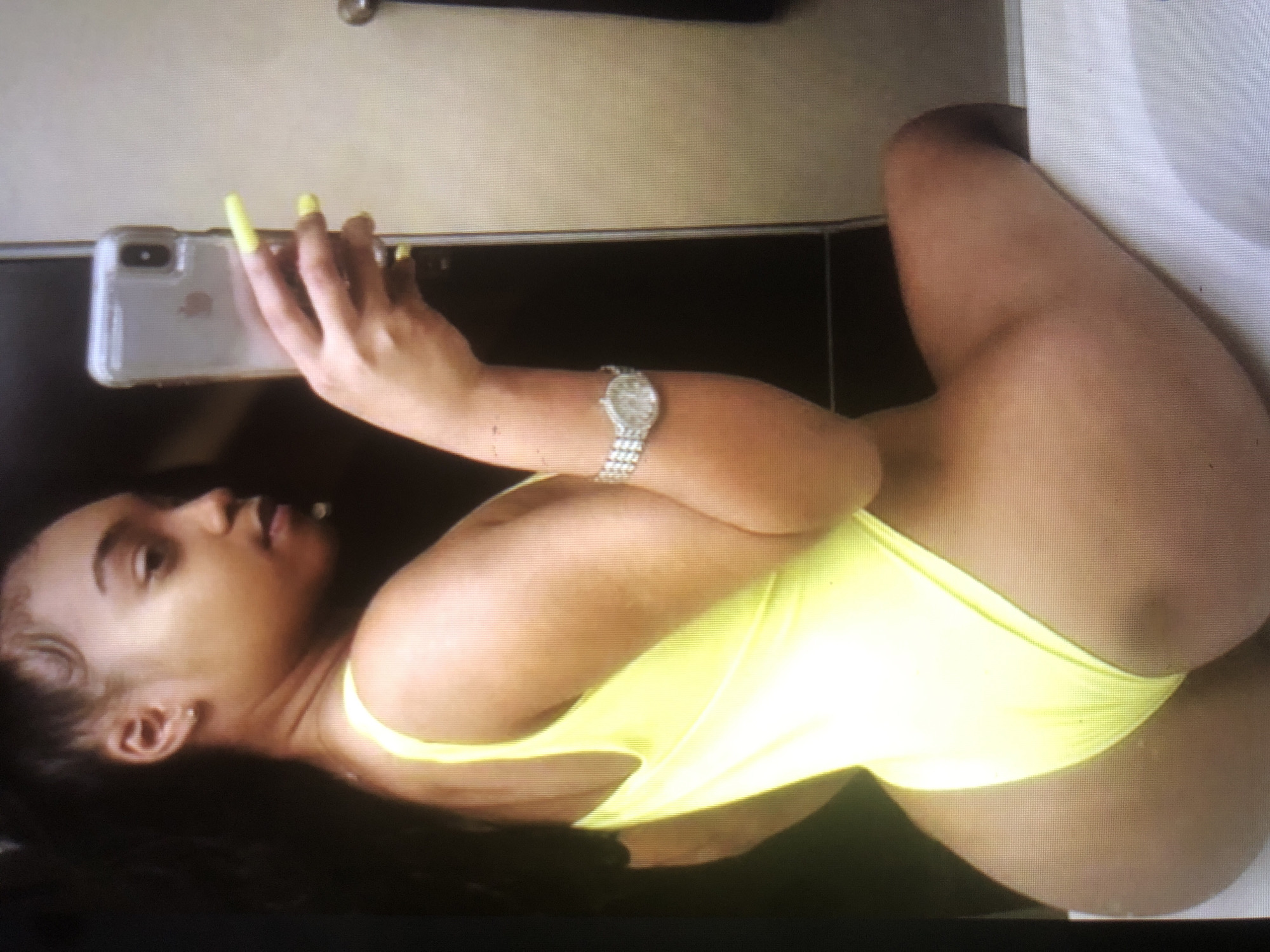 The media file shows a woman in a bright yellow swimsuit, holding a smartphone. She is lying down, with her head tilted back and her gaze directed at the phone. Her fingernails are painted a bright yellow, matching her swimsuit. She is wearing a silver watch on her left wrist. The background appears to be a tiled wall. The image is oriented vertically, suggesting it might be a portrait or a selfie. The lighting suggests an indoor setting, possibly a bathroom or a changing room, with bright overhead lighting. No specific time of day or weather conditions can be determined. There is no visible text in the image.