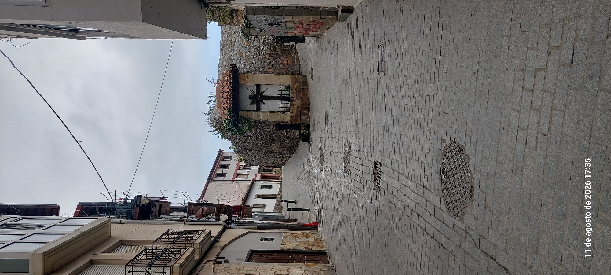 This is a full-frame, eye-level, vertical shot of a cobblestone street in Castro-Urdiales, Spain, on a cloudy day. The street slopes gently uphill and appears to fork in the distance. On the left, a building with light-colored stone and dark wood accents, featuring balconies with wrought-iron railings, lines the street. Further up on the left, another building with white walls and red trim is visible. On the right side of the street, a rustic stone wall is prominent, with a small, sheltered alcove containing a dark wooden cross. A fountain is also visible near the base of the wall. The street is paved with gray cobblestones and has several manhole covers and drain grates. In the foreground, a large, circular manhole cover with a patterned surface is visible. The sky is overcast. The timestamp "11 de agosto de 2026 17:35" is superimposed on the bottom left corner of the image.