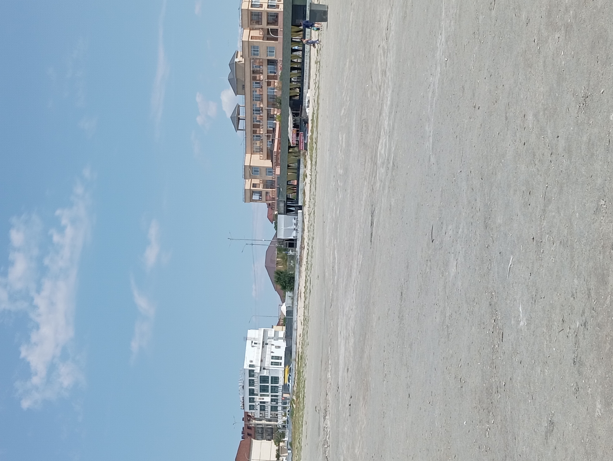 In Ömirzaq, Kazakhstan, a wide, sandy beach stretches towards a light blue sky with scattered white clouds. In the mid-ground, several buildings line the shoreline. To the left, a modern, multi-story white building with glass windows stands prominently, displaying signage that appears to read "URBAN HOUS-SPA". Adjacent to it, a red-tiled roof building is visible. Further to the right, a long, low building with a green roof and open seating areas with yellow curtains extends along the beach. Behind this, a larger, peach-colored hotel building with multiple balconies and a flat roof with a central structure dominates the background. Two individuals, one an adult and a child, are walking on the sand towards the right side of the image, near a dark gray trash receptacle. The overall scene depicts a coastal resort area on a clear day.