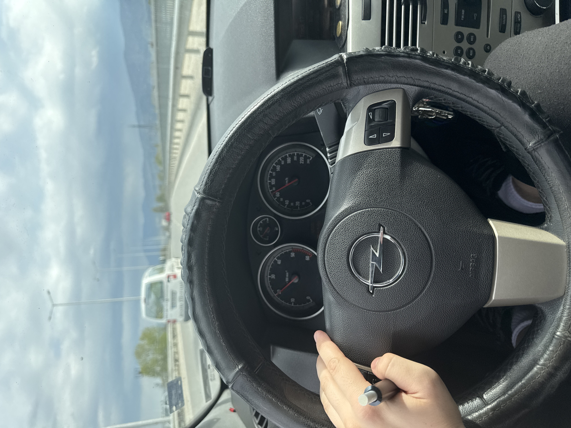 The image shows the interior of a car, specifically the steering wheel with an Opel logo. The steering wheel has an aftermarket cover. The car is in motion on a road in what appears to be an urban area. Through the windshield, one can see other vehicles, street lights, and a cloudy sky. Given the Opel logo on the steering wheel, the scene is likely in Europe. Without further information, I cannot confirm that the location is Plovdiv, Bulgaria. A person's hand is partially visible on the steering wheel, holding a pen-like object. There's another person's sock visible in the top right of the steering wheel. The car's dashboard gauges are visible behind the steering wheel. A car key is dangling above the steering wheel.