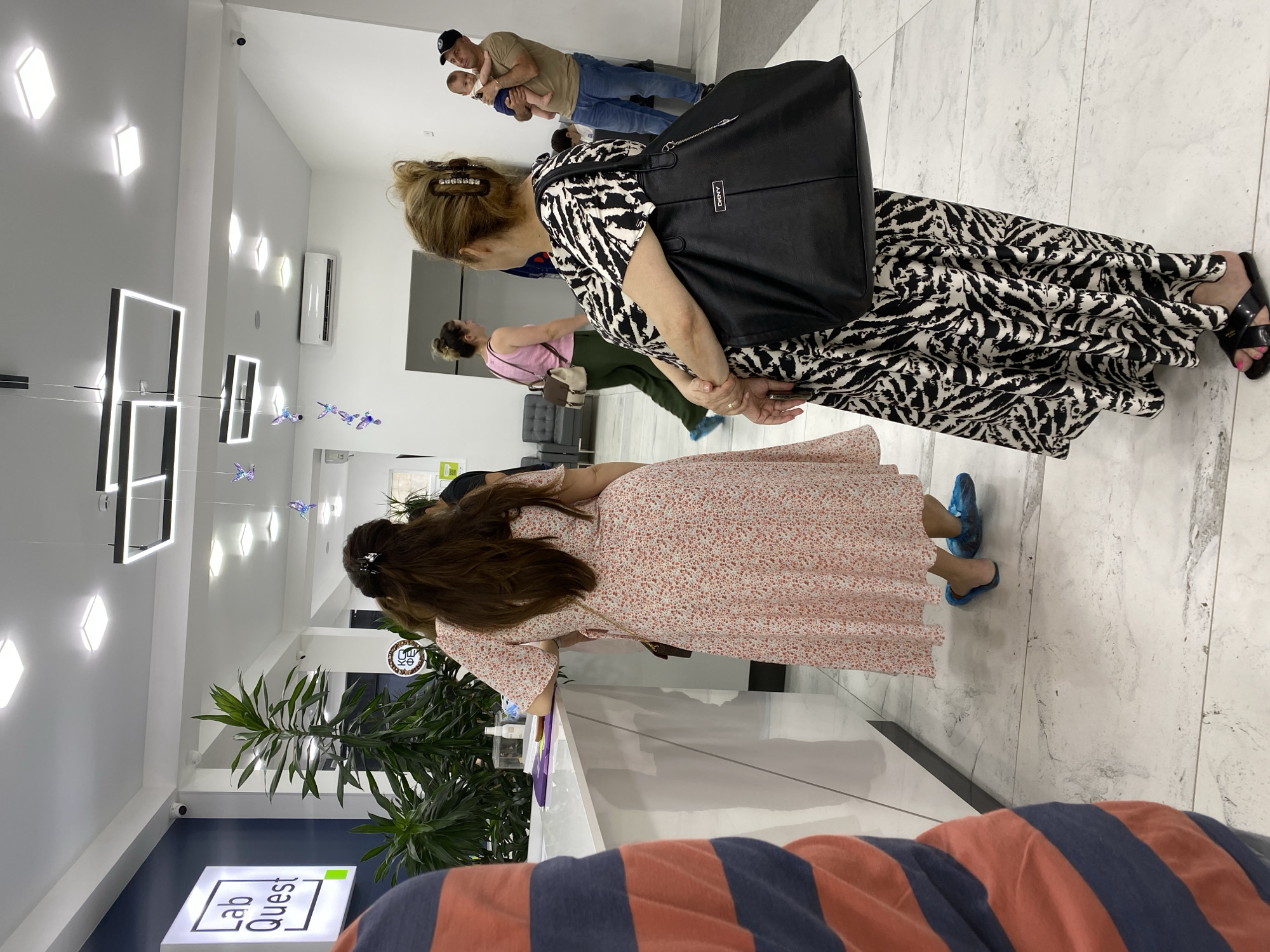 The image depicts the interior of a modern laboratory clinic, specifically "Lab Quest" in Sokhumi, Abkhazia. The scene shows several people in a waiting area or at a reception counter. In the foreground, a person wearing a striped shirt is partially visible. To their right, a woman in a floral dress stands at a white counter, appearing to interact with staff or fill out paperwork. Further back, a woman in a black and white zebra-print dress holds a large black tote bag. To her right, a man in a light brown t-shirt and blue jeans holds a baby. Another woman in a pink top and green pants walks in the background. The clinic features bright, white lighting, sleek flooring, and decorative elements like hanging butterfly figures. Plants are visible near the entrance and reception area. An air conditioning unit is mounted high on a wall. The overall impression is one of a clean, contemporary healthcare facility.
