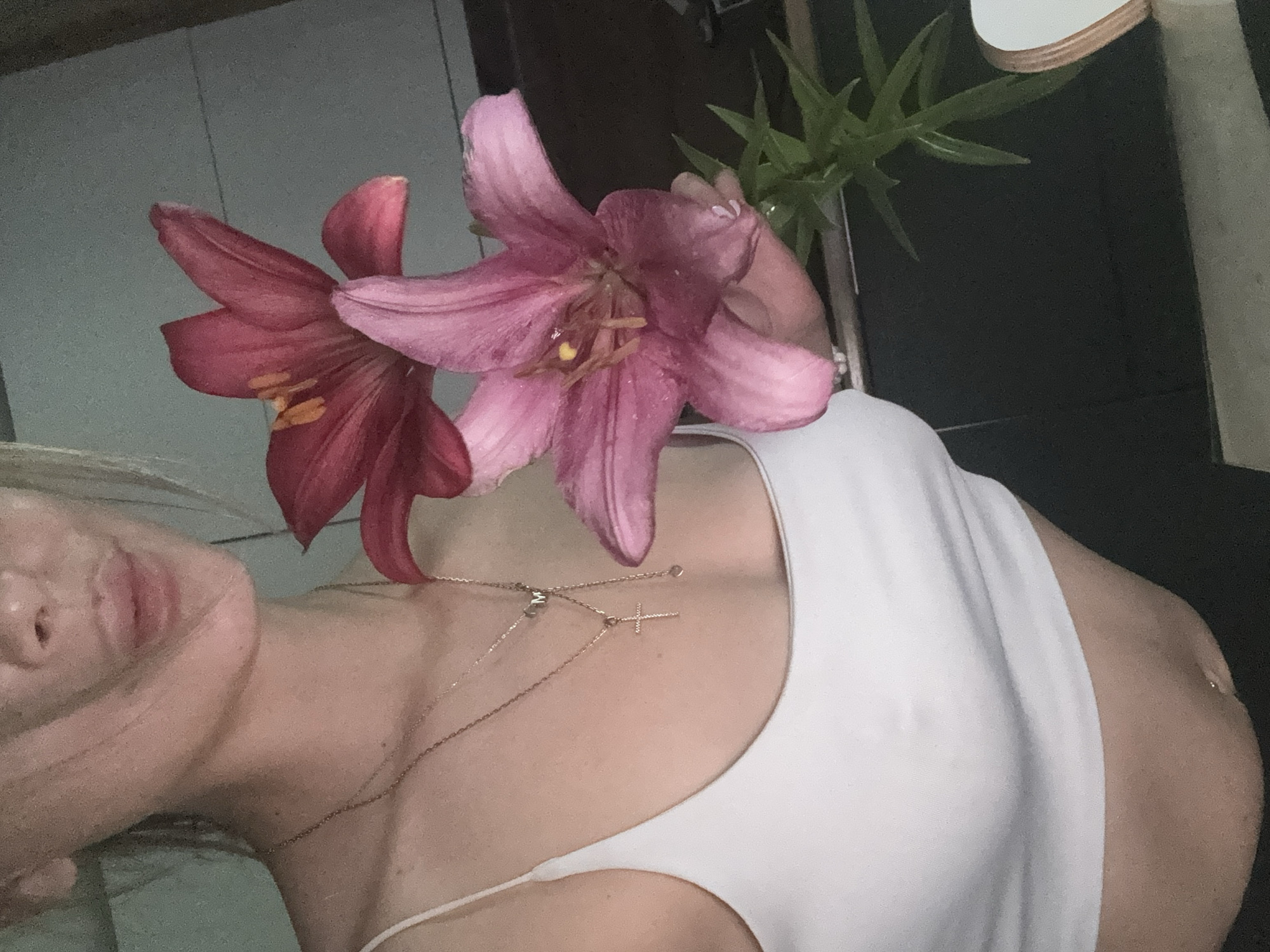 The image shows a close-up of a person holding two pink lilies. The person is wearing a white tank top and several necklaces, including one with a cross pendant and another with the letter "M". The background is dark and out of focus, suggesting an indoor setting. There is no visible indication of the location being in Tula, Russia.