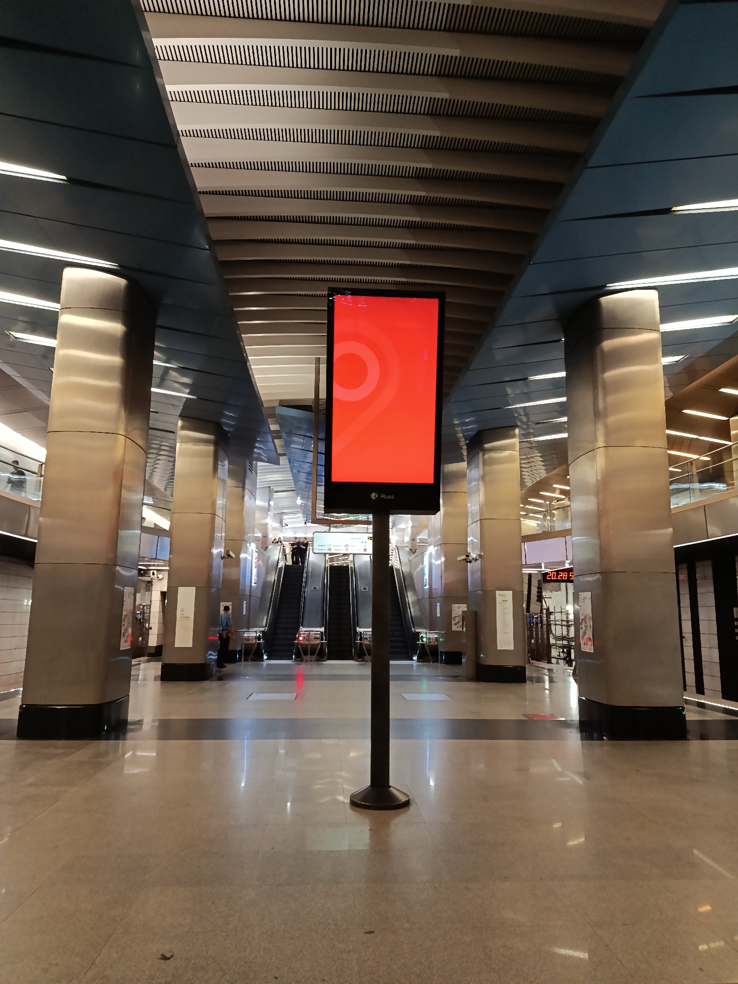 The image shows the interior of a subway station in Moscow, Russia. The station features a modern design with large, cylindrical, metal-clad columns supporting the ceiling. The ceiling has a distinct pattern of parallel lines. In the center of the frame is a digital advertising display with a red screen. Behind the display, escalators lead to another level of the station. People can be seen using the escalators and walking in the background. The floor is made of polished stone tiles, reflecting light. The scene appears clean and well-maintained.