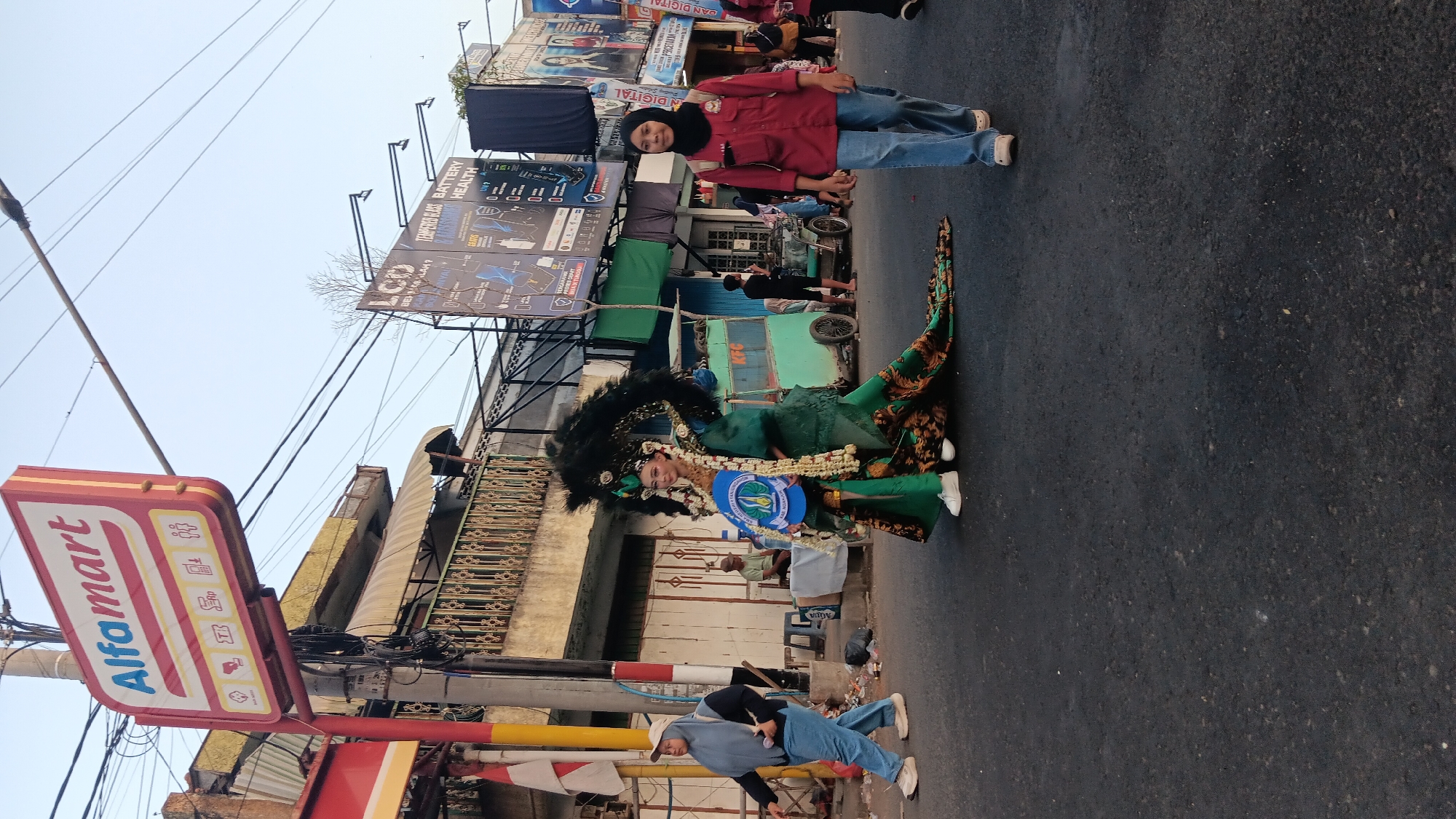 In Tanjung Anom, Indonesia, a street scene unfolds with a person in elaborate green costume and a large headdress walking down the asphalt road. The costume features intricate patterns and a long train. To the left, a woman in a hijab and jeans walks away from the camera. To the right, a woman in a red shirt and jeans walks towards the camera, also wearing a hijab. In the background, an Alfamart sign is visible, along with buildings, power lines, and a green food cart. The sky is clear.
