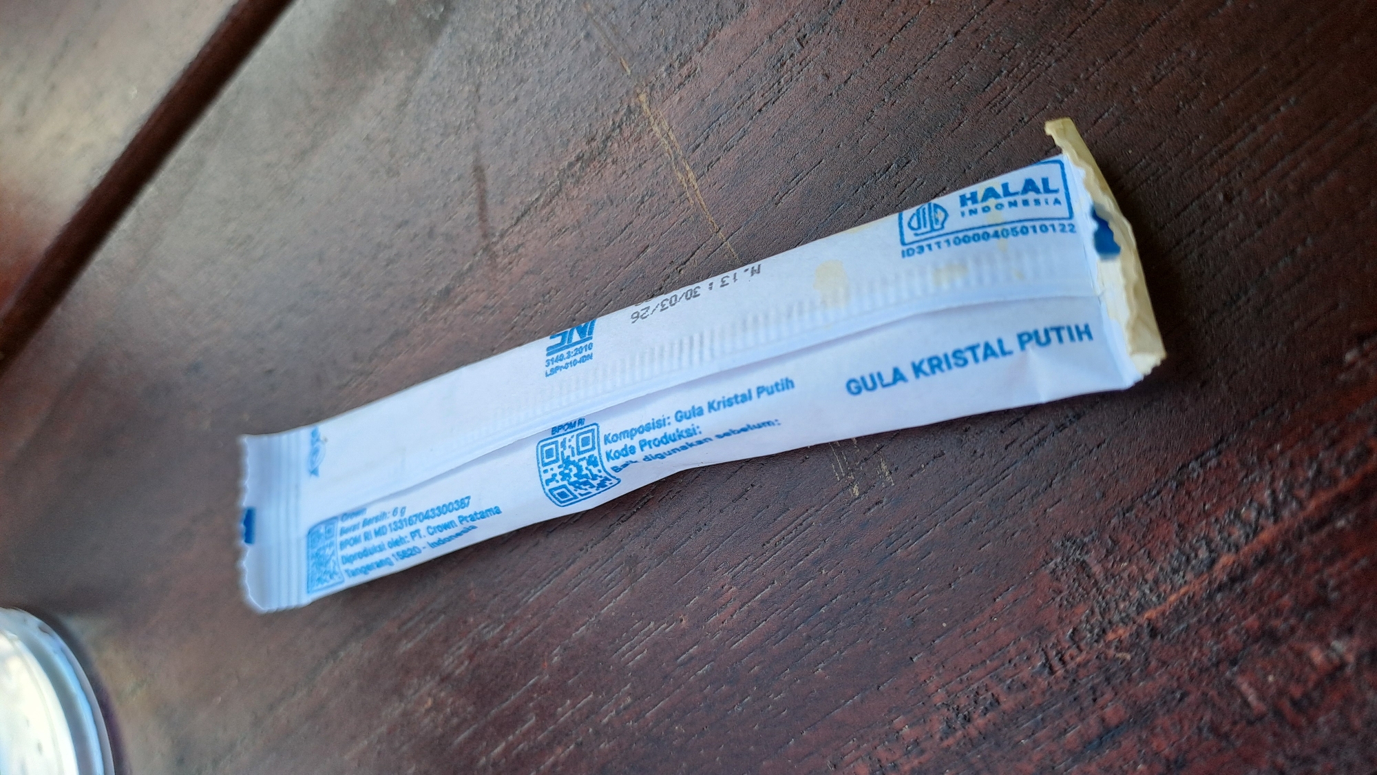 A sugar packet is placed on a wooden surface. The packet is white with blue text and graphics. The text on the packet is in Indonesian and includes information such as "GULA KRISTAL PUTIH" (White Crystal Sugar), "Komposisi: Gula Kristal Putih" (Composition: White Crystal Sugar), and a Halal certification logo. There are also QR codes printed on the packet. The wooden surface appears to be a table with a visible wood grain. The lighting suggests an outdoor or well-lit indoor environment. The image is taken from a slightly elevated angle, focusing on the sugar packet. The specific location of Bireuenmeunasahdayah, Indonesia is not visually discernible from the image itself; this information would be external context.