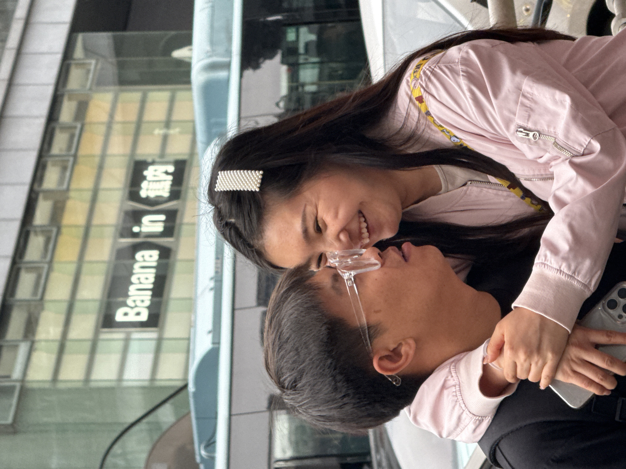 A woman with long dark hair, adorned with a pearl barrette, smiles while hugging a man wearing clear-framed glasses and a light pink bomber jacket over a black shirt. She has her arms wrapped around his neck, and her hands are holding his. He looks upwards. A building with the sign "Banana in" is visible in the background, hinting at the location in Liangjiang, China. The setting appears to be an outdoor urban environment, likely near a bus or building with large windows.