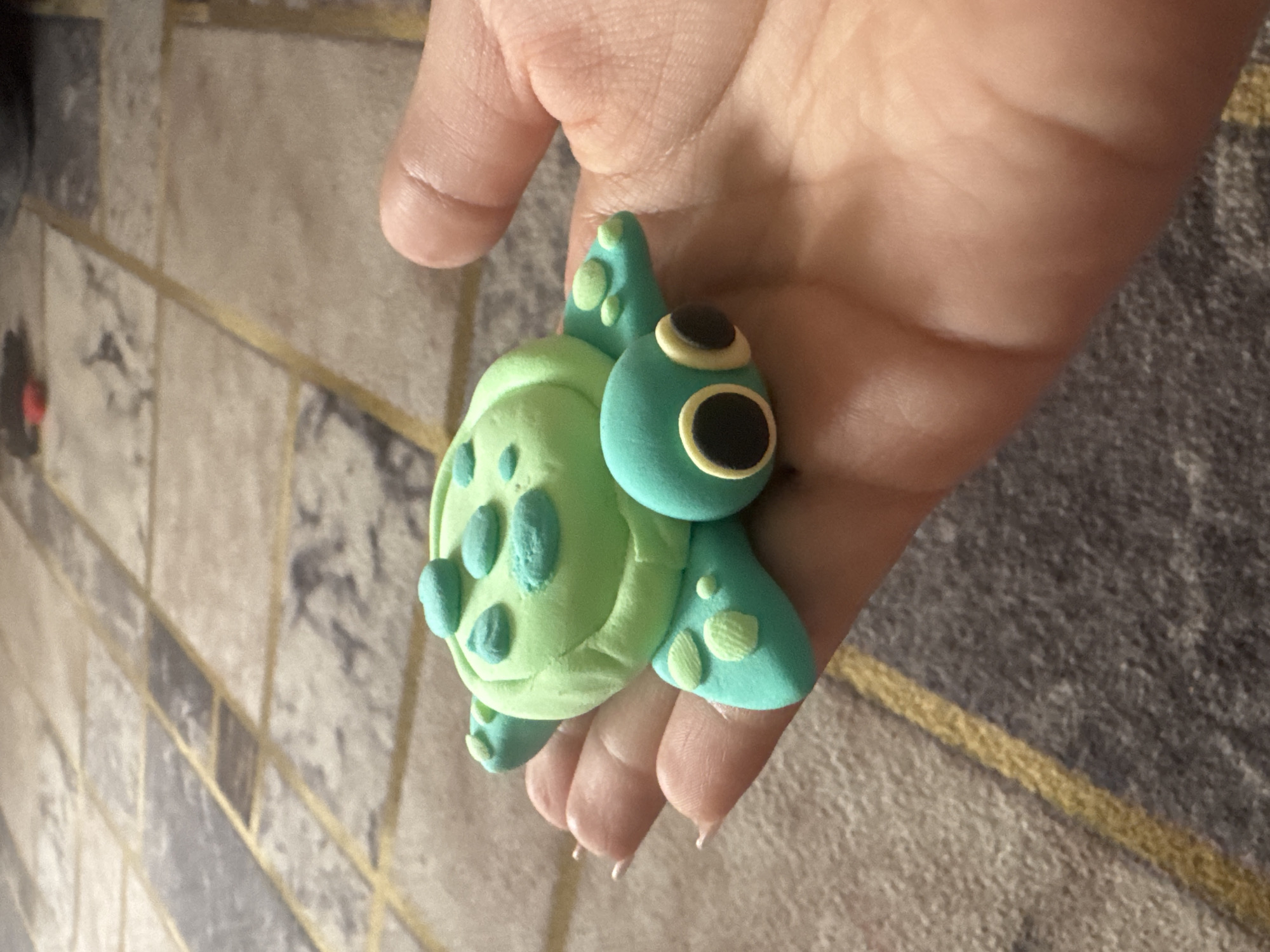 A close-up shot captures a light-skinned hand holding a small, handcrafted turtle figure. The hand, likely an adult's, has a gentle grip on the toy, with fingers slightly curled. The turtle is made of what appears to be clay or play-doh, featuring a two-toned green color scheme. Its shell is a lighter lime green with several raised, darker green spots, while its head, four flippers, and small tail are a more vibrant teal. The teal parts also have lighter green accents, some with textured patterns. The turtle's head is round, showcasing large, expressive eyes with black pupils encircled by cream-colored irises, giving it a friendly, cartoonish appearance.
The background shows an indoor setting, likely a floor or a large rug, characterized by a geometric pattern. This pattern includes varying shades of gray, from light to dark, interspersed with thin, intersecting gold lines, creating a tiled or interwoven design. The lighting is ambient and typical of an indoor environment, offering no specific cues about the time of day or weather. No text is visible in the image. The scene conveys the act of presenting or admiring a small, handmade craft, situated in an indoor location.