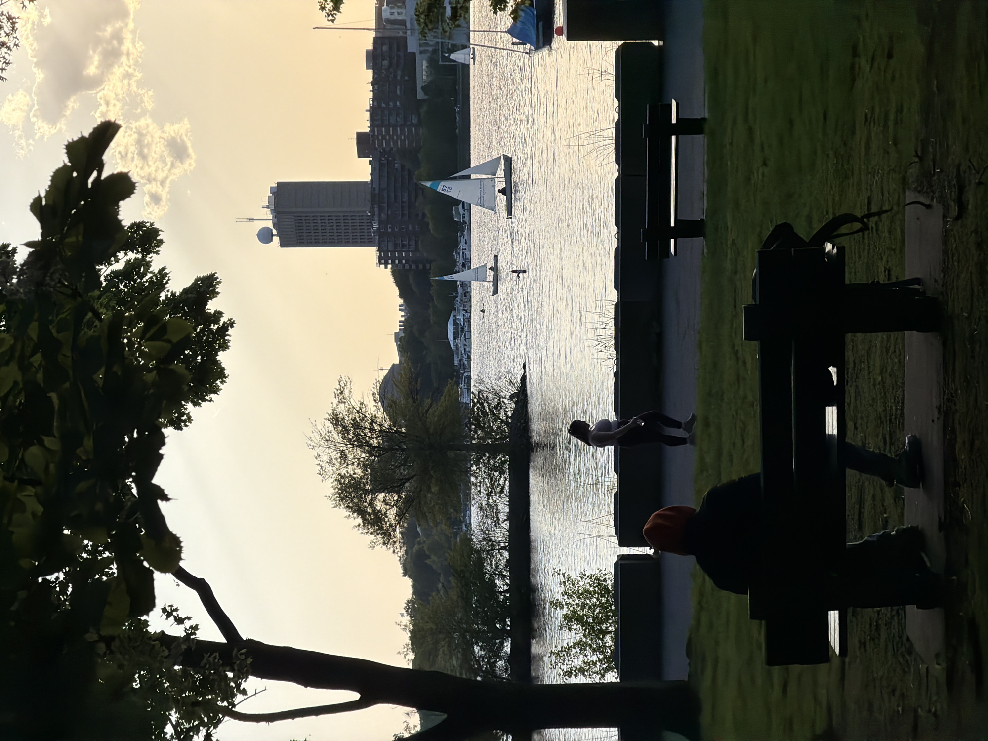 The image shows a view of the Charles River in Boston, United States of America, from a park bench. On the right, a person is sitting on the park bench, and another person is leaning on the edge near the water. Sailboats are on the river, and buildings are visible in the background, including a tall building with a distinctive round structure on top. The sky is bright, indicating sunset or sunrise. Trees frame the left side of the image.