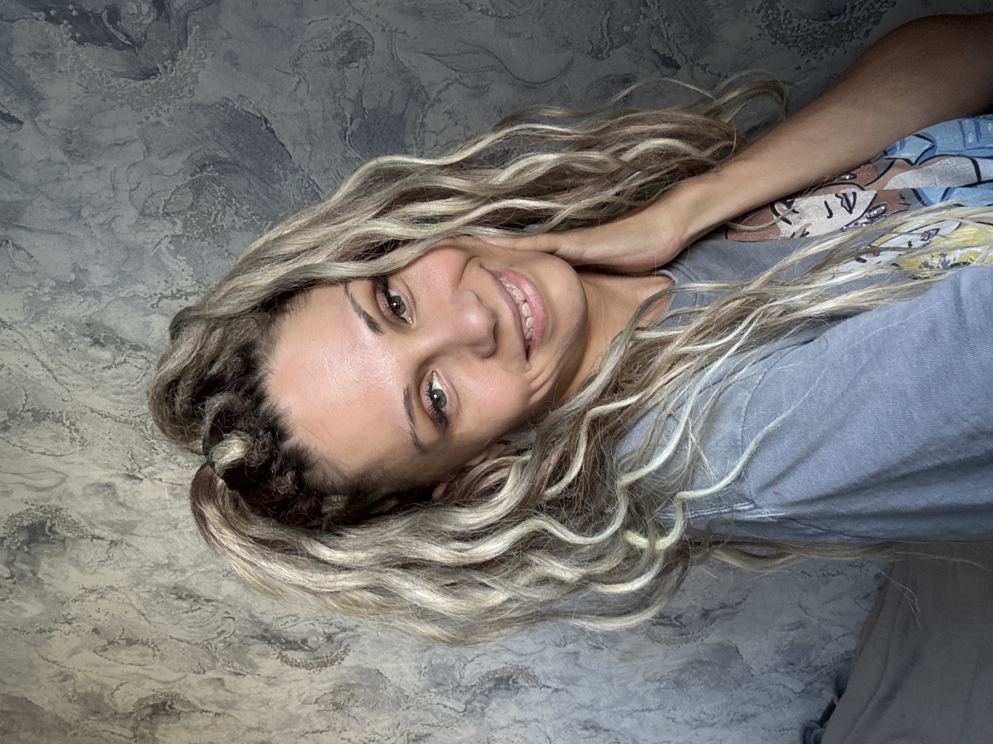 A woman with blonde, curly hair styled in a high ponytail, is shown from the chest up, smiling and touching her cheek with her right hand. She is wearing a grey t-shirt with a graphic print of three illustrated women. The background is a textured grey wall. The image appears to be taken indoors. There is no indication of the location being the city of Rusanovo, Russia.
