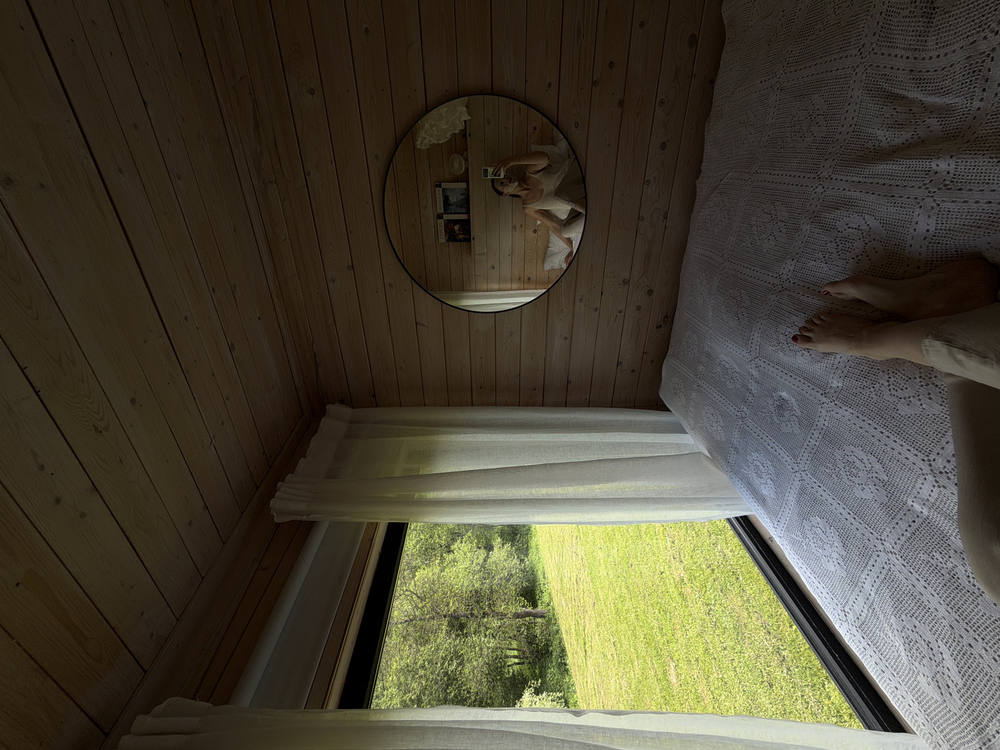The image shows the interior of a room with wooden walls, likely a cabin or small house. A triangular window with sheer white curtains reveals a view of a grassy field and trees outside, suggesting a rural or natural setting. On the right side of the frame is a bed covered with a white crocheted bedspread. A person's legs and feet are visible on the bed, with red nail polish on the toes. A round mirror hangs on the wall, reflecting a person taking a photo with a smartphone. Behind the person in the mirror, there are posters on the wall. There is no evidence to suggest the location is Prudki, Russia.