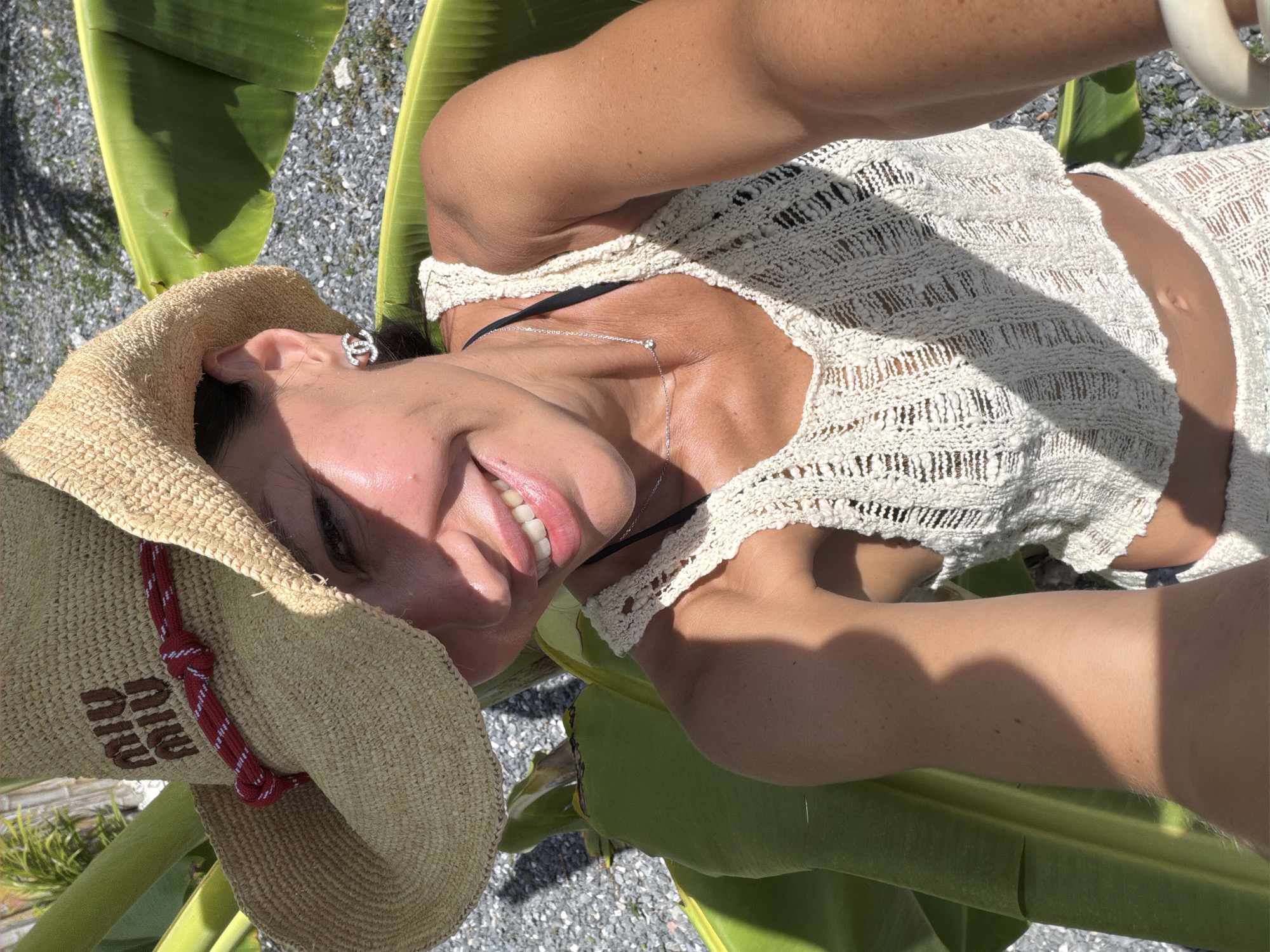 A woman is pictured outdoors in Baan Bo Phut, Thailand. She is wearing a straw cowboy hat with a red and white rope band, a white crocheted tank top, and a matching white crocheted skirt or shorts. She has on a diamond pendant necklace and Chanel earrings. The woman is smiling and looking at the camera. She is surrounded by large green banana leaves, and the ground is covered in small gray stones. The lighting suggests it is a sunny day.