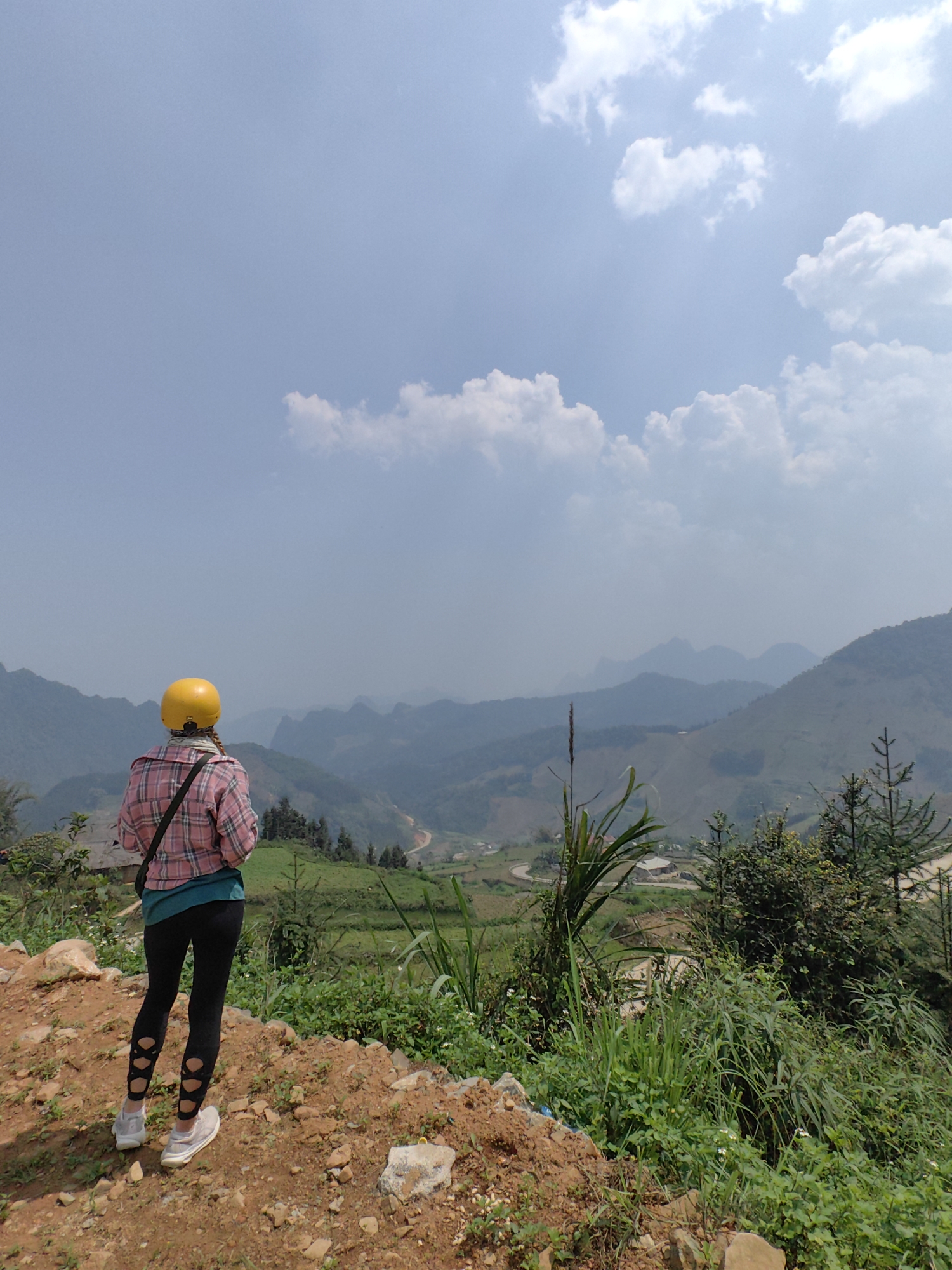 A woman wearing a yellow helmet and a pink plaid shirt stands on a dirt patch, looking out at a mountainous landscape in Xã Cần Yên, Vietnam. She has black leggings with cutouts at the calves and light-colored sneakers. The landscape features green hills, some with terraced fields, and distant mountains shrouded in haze. The sky is blue with scattered white clouds. Green vegetation including grasses and small trees is in the foreground.