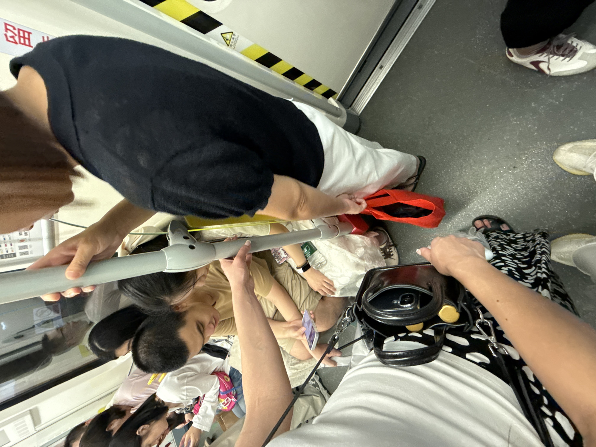In Haizhu District, China, inside a crowded public transport vehicle, a person wearing a black top and white pants is holding a red bag while standing near a door. Other passengers are visible, some seated and others standing. One person is holding a smartphone, and a black handbag with gold accents is resting on the floor. The environment appears to be a train or subway carriage, indicated by the pole, seats, and warning signs on the door. The lighting suggests an indoor, artificial environment.