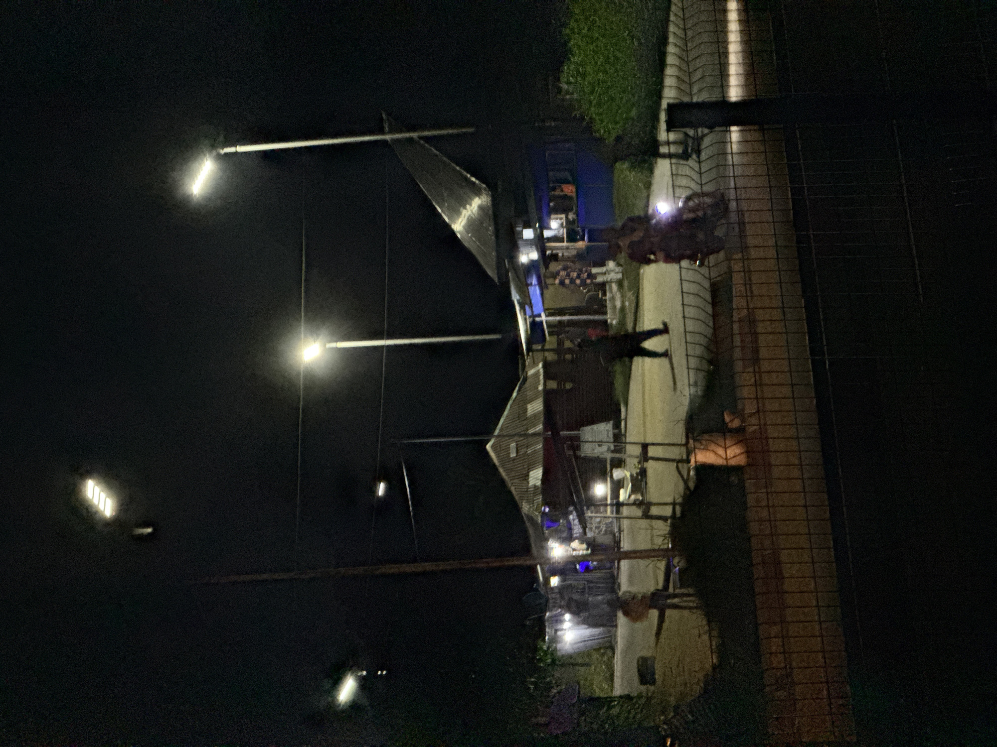 The image depicts a night scene on a street in Uyo, Nigeria. The street is illuminated by several streetlights. In the foreground, a wire fence runs horizontally across the frame. Beyond the fence, a paved area is visible with several structures, including a stall with a corrugated metal roof and another with a blue awning.
Several individuals are present. Two people are seen riding a motorcycle with its headlight on, moving from left to right in the mid-ground. To the left of the motorcycle, a person stands near a wooden pole and a cart. Further back, other figures are visible near the stalls. A person in a plaid shirt is standing in front of one of the stalls. A single person is seen walking from left to right in the mid-ground to the left of the motorcycle. The ground appears to be a mix of pavement and dirt. Green foliage is visible on the right side of the image. The overall atmosphere is that of a busy street or market area at night.