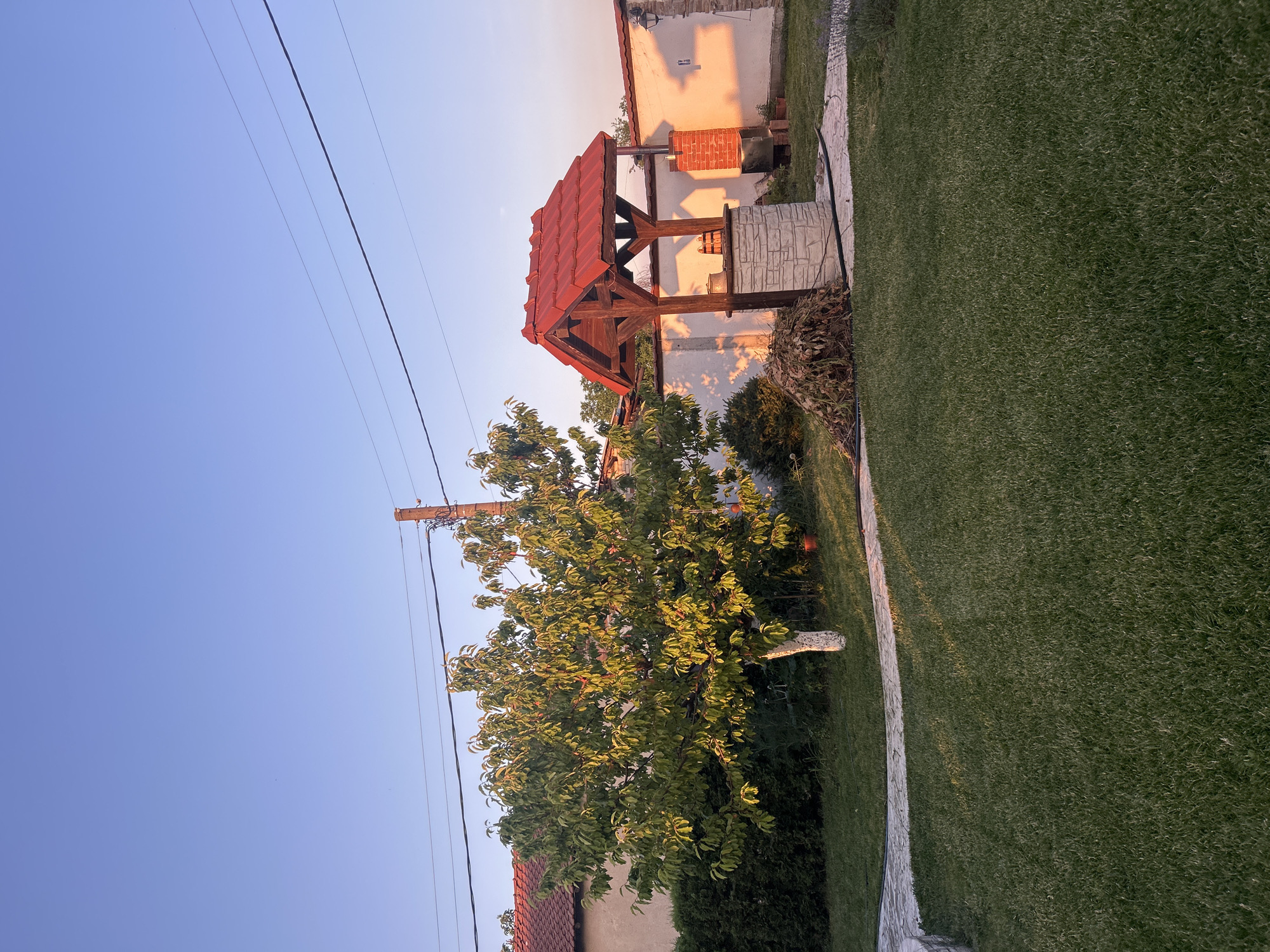 In the city of Voyvodino, Bulgaria, an outdoor scene unfolds. The image captures a residential yard with a well-maintained lawn. To the left, a mature tree with green leaves stands, its trunk painted white. Behind the tree, a building with a red-tiled roof is visible, along with a dark green hedge. In the center right, a picturesque well with a red-tiled roof structure is present, made of stone and wood. Adjacent to the well, a pile of dried leaves and branches rests on the ground. A brick chimney structure stands to the right of the well. A paved pathway curves through the lawn, leading towards the well. The sky above is a clear, gradient blue, transitioning to a warmer hue near the horizon, suggesting either sunrise or sunset. Overhead, electrical wires are strung between poles, one of which is visible on the right side of the frame. The scene is bathed in a warm, soft light, characteristic of the golden hour.