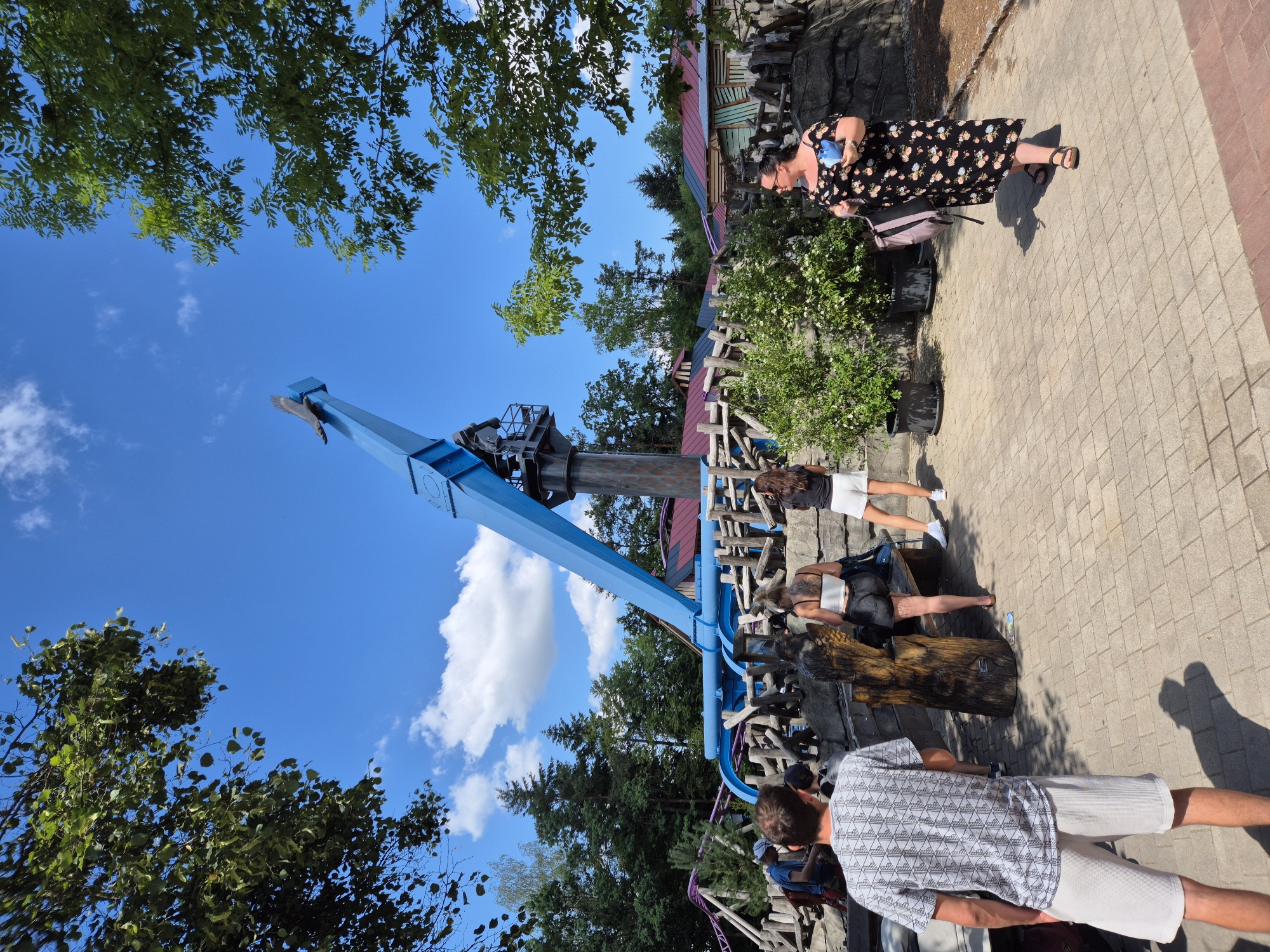 The image shows a sunny day at an amusement park. In the background, a large blue amusement park ride dominates the frame, with a statue of a bird perched on top. The ride appears to be a drop tower or a similar thrill ride. In the middle ground, a wooden fence encloses an area with more amusement park structures, including a roller coaster track with purple rails. In the foreground, several people are walking on a paved pathway. A man in a patterned t-shirt and white shorts is walking away from the camera. To his left, a woman in a black floral dress is walking towards the camera, carrying a bag and a blue object. Other people are visible in the background, near the attraction and the fence. The park is surrounded by trees, indicating a natural setting. The overall atmosphere is that of a lively outdoor recreational space. The scene is located in the city of Reisbach, Germany.
