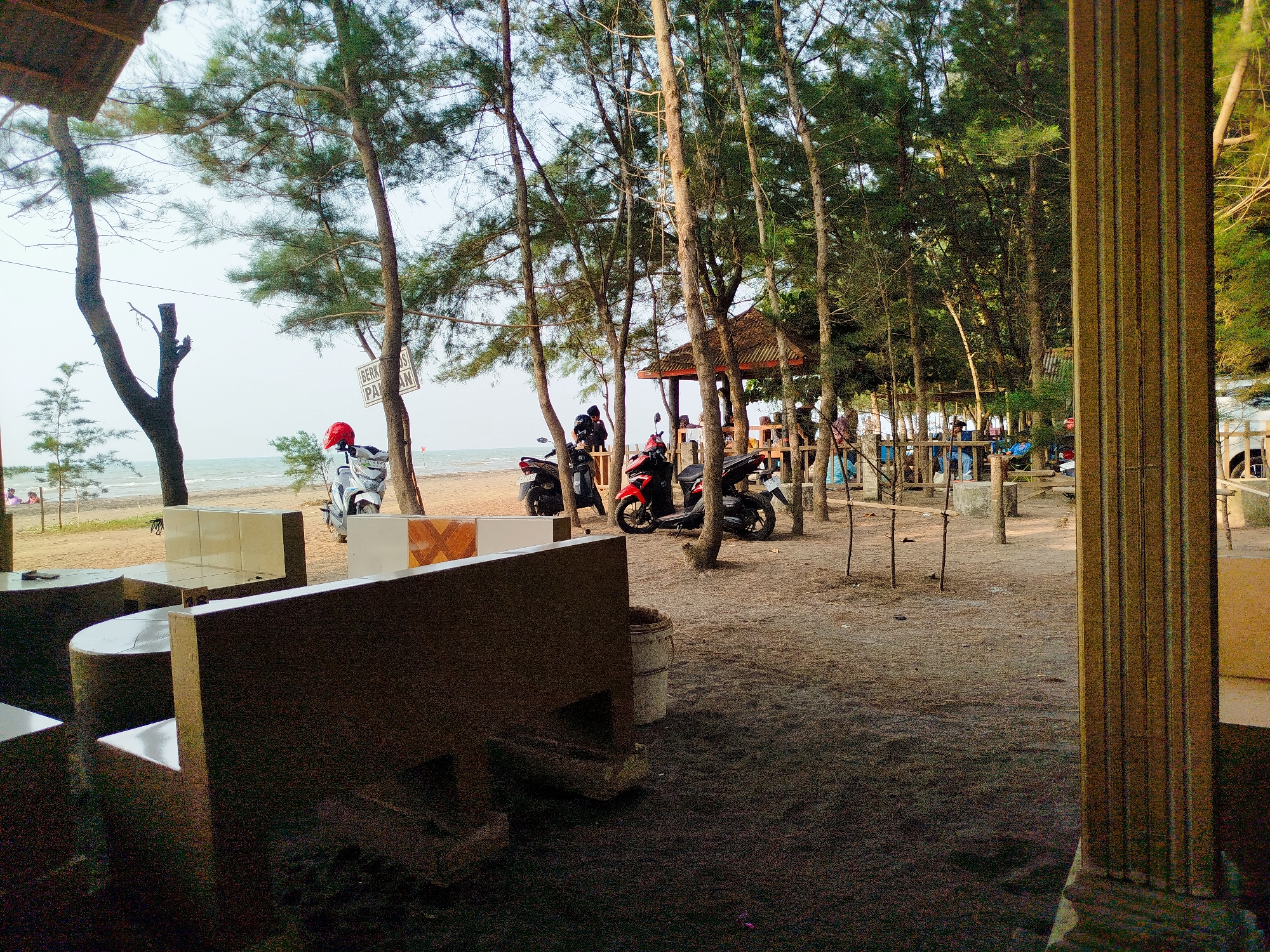 This image depicts a seaside area in Jungsemi, Indonesia, likely a restaurant or rest area with outdoor seating. In the foreground, there are concrete benches and tables. Beyond the seating area, there are several trees lining a sandy path leading towards the beach. In the middle ground, a small hut or gazebo is visible, surrounded by more trees. Several motorcycles are parked in the area, suggesting a mode of transportation for visitors. The beach and the ocean are visible in the background under a cloudy sky. A sign with text, partially obscured by trees, is also present. The overall scene suggests a casual, outdoor recreational space near the coast.