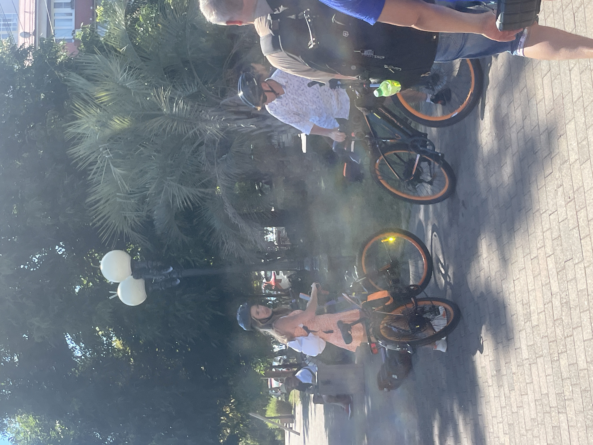 The image shows a sunny day scene in Sochi, Russia, with people on bicycles. In the foreground, a woman with blonde hair, wearing a peach-colored dress and a black helmet, is on a black bicycle with orange rims. She is looking to her left and holding the handlebars. Next to her, a man wearing a white patterned shirt and sunglasses, also with a black helmet, is on another black bicycle with orange rims, also looking to his left. Behind them, a person's back is visible, wearing a backpack and denim shorts, facing away from the camera. Further in the background, another person is sitting on a bench. The setting appears to be a park or promenade with trees and palm trees, and a building visible in the distance. The ground is paved with brick-like tiles. There is a lamppost with two white globes visible in the middle ground.