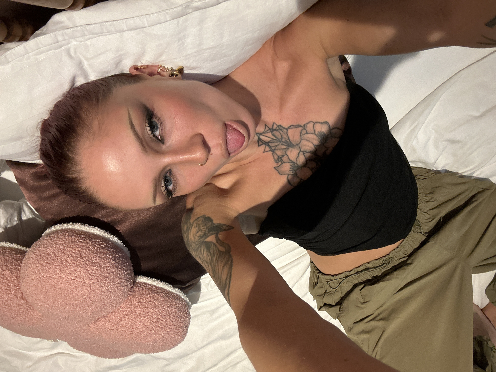 A woman lies on a white bed, propped up by pillows. She has purple hair with dark roots, and her eyes are bright blue. She is wearing a black tube top and olive green cargo pants. She has tattoos on her chest and arm, as well as piercings in her ears and nose. A hand, presumably belonging to another person, is visible on her arm. The room is dimly lit, suggesting it might be nighttime. The setting appears to be a bedroom. The woman's expression is playful and slightly suggestive, with her tongue sticking out. The overall mood is intimate and casual. The image seems to have been taken indoors, and there are no visible clues about the weather or time of day beyond the lighting. There is no visible text in the image.