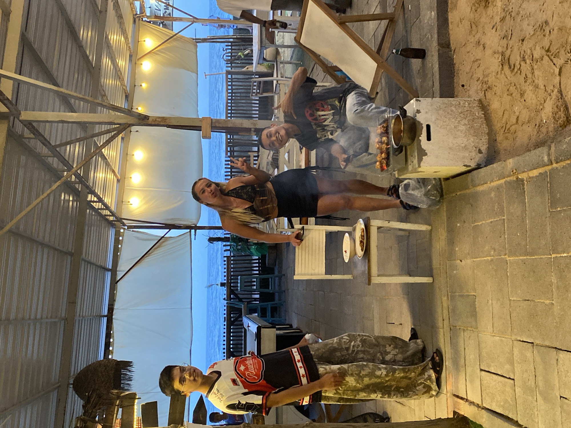Three young adults are posing outdoors at what appears to be a beachfront restaurant in Mũi Né, Vietnam. The sky is a clear, deep blue, suggesting it is dusk or early evening. The background features the ocean and a wooden railing. The foreground is a paved area with some sand.
On the left, a young man wearing glasses, a black, white, and red jersey, and camouflage pants stands holding a bottle. He is looking at the camera with a slight smile.
In the center, a young woman with blonde hair is striking a peace sign with her right hand and looking towards the camera. She is wearing a dark, patterned halter top and a black skirt.
To the right of the woman and slightly behind her, a young man is sitting down. He is wearing a dark t-shirt with a graphic design and light-colored jeans. He is smiling and making a "rock on" hand gesture. In front of him is a small grill with skewers of meat cooking. Next to the grill is a pot on a white concrete block.
The overall atmosphere is casual and social, with the group appearing to be enjoying a meal or preparing one. The lighting suggests artificial lights are also in use, likely from the restaurant's overhead structure.